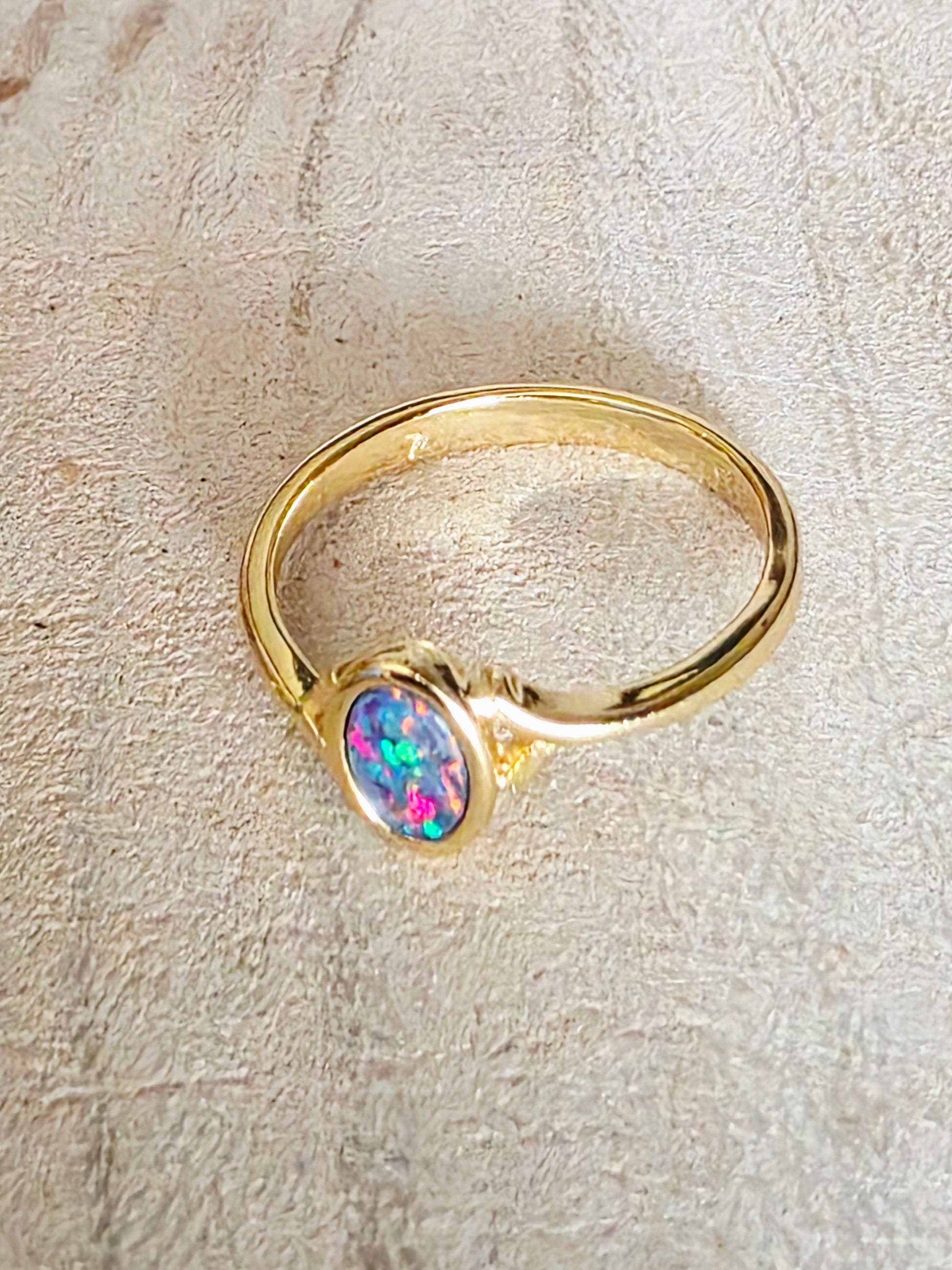 A gold ring featuring an oval-cut iridescent opal stone with vibrant pink, blue, and green flashes, set against a textured beige background.
