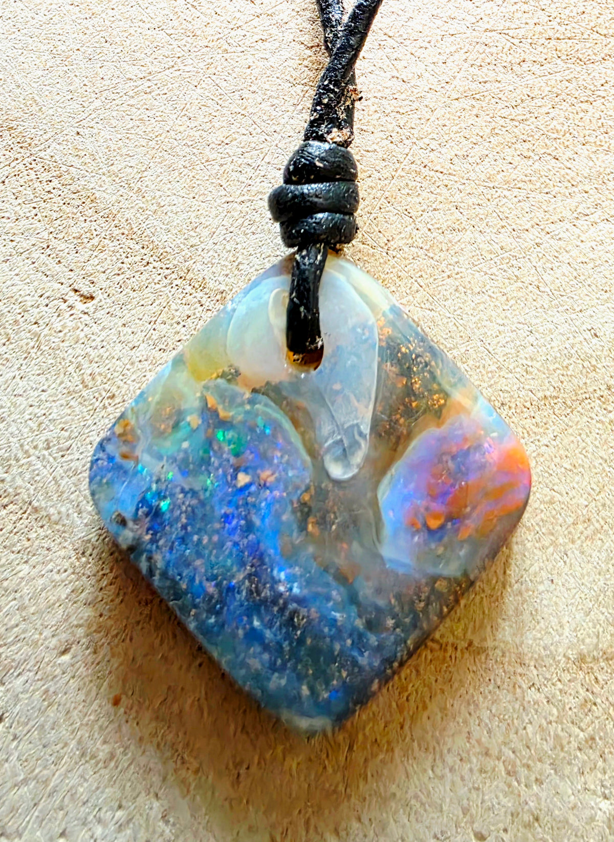 A square opal pendant necklace with a black cord, featuring vibrant blue, green, orange, and white play-of-colour opal, displayed on a textured beige surface.