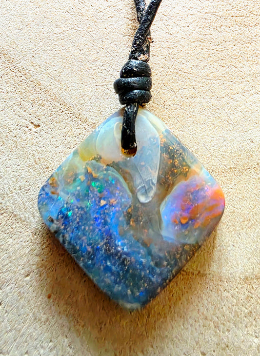 A square opal pendant necklace with a black cord, featuring vibrant blue, green, orange, and white play-of-colour opal, displayed on a textured beige surface.