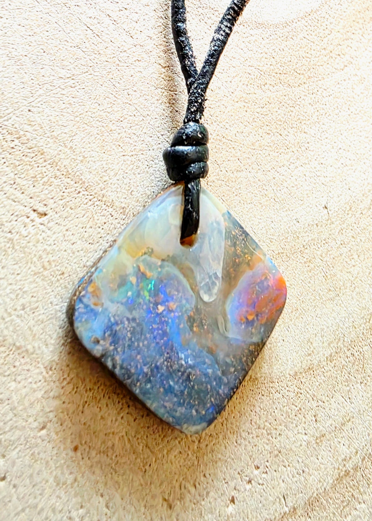 A square-shaped opal pendant with a black cord, displayed on a light wood background.