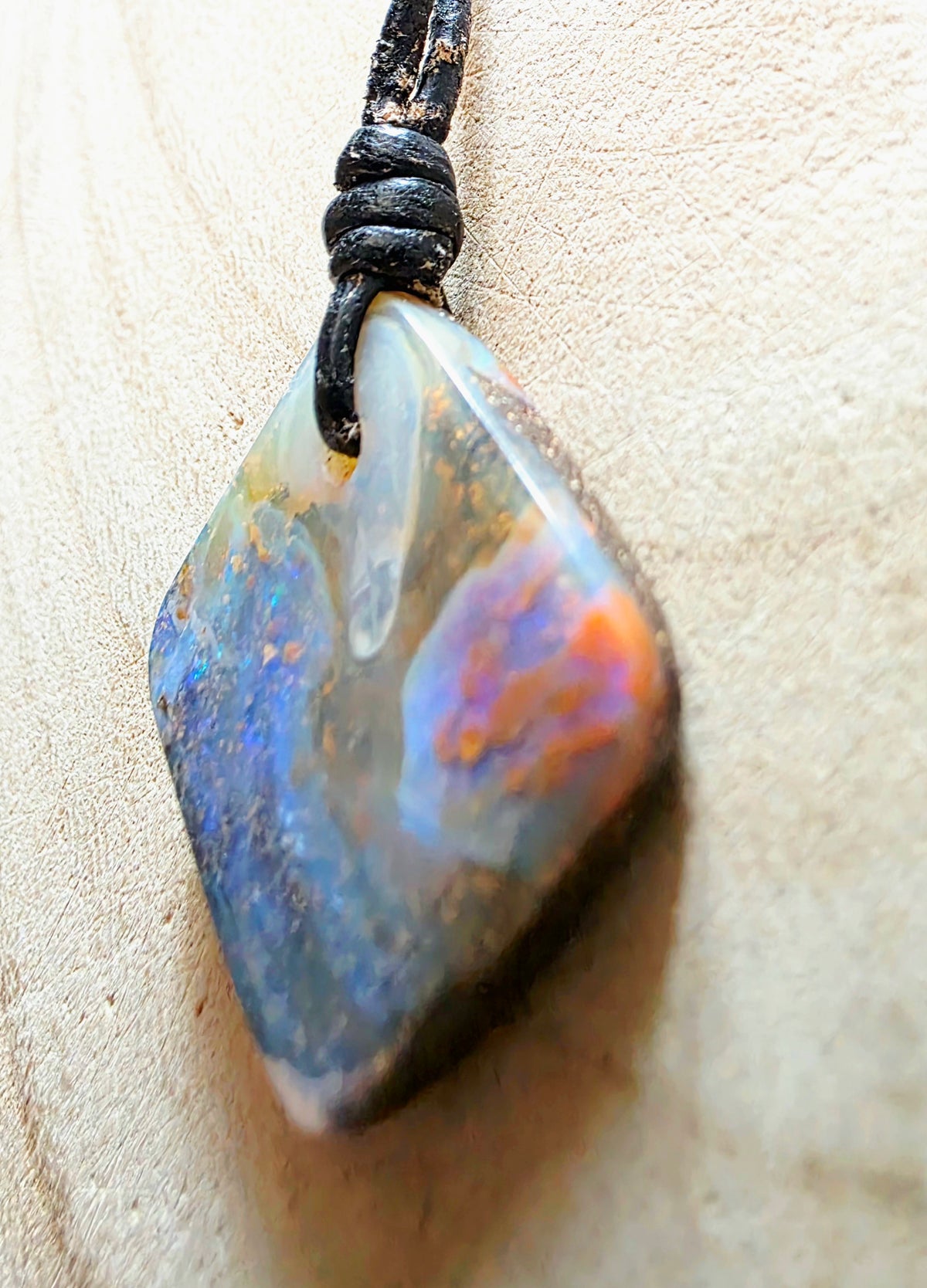 A natural opal pendant with iridescent blue, orange, and purple flashes, suspended from a black cord on a light beige textured background.