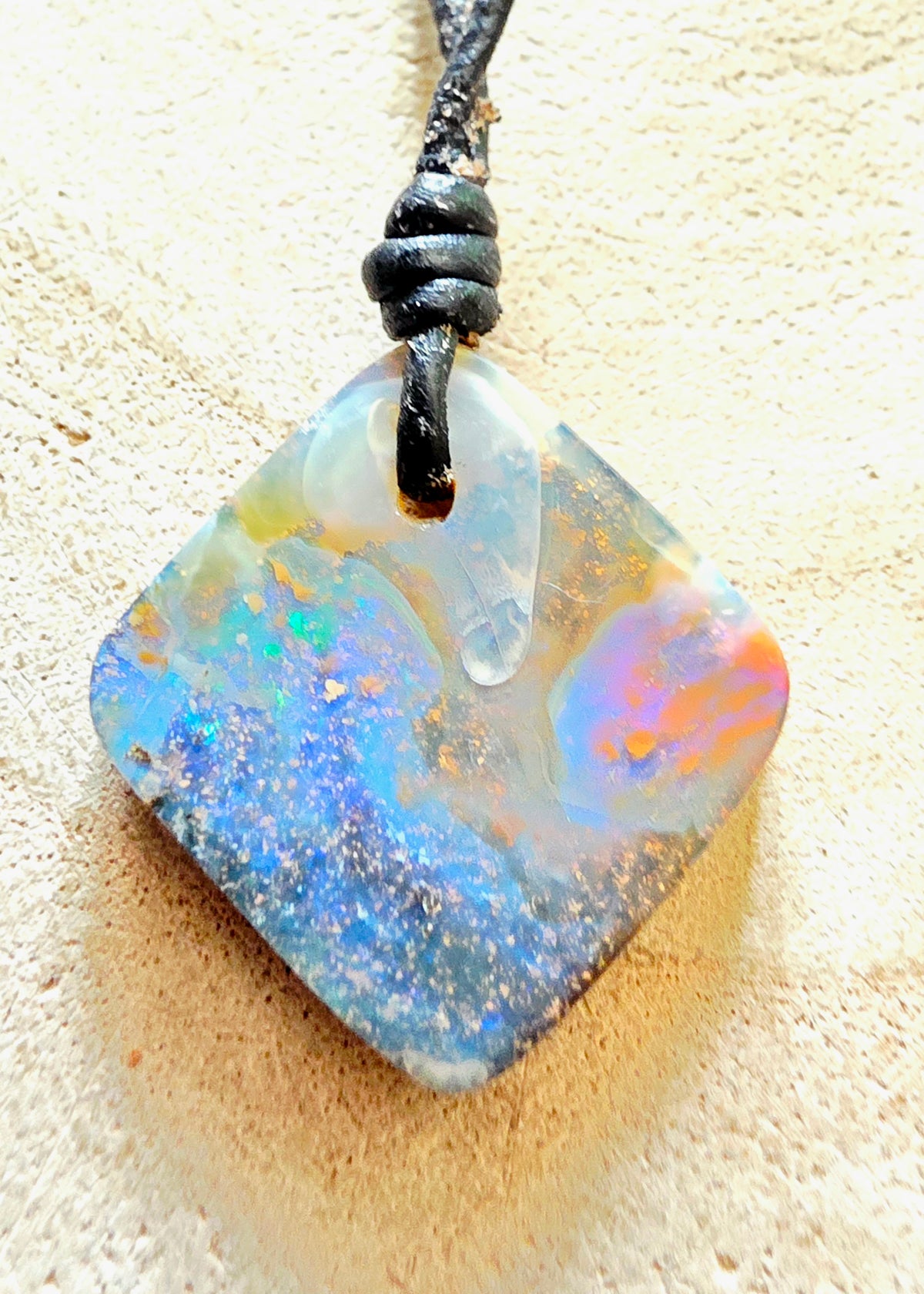 A square opal pendant necklace with a black cord, resting on a textured beige surface. The opal displays a vibrant play-of-colour with blue, green, orange, and white iridescent patterns.