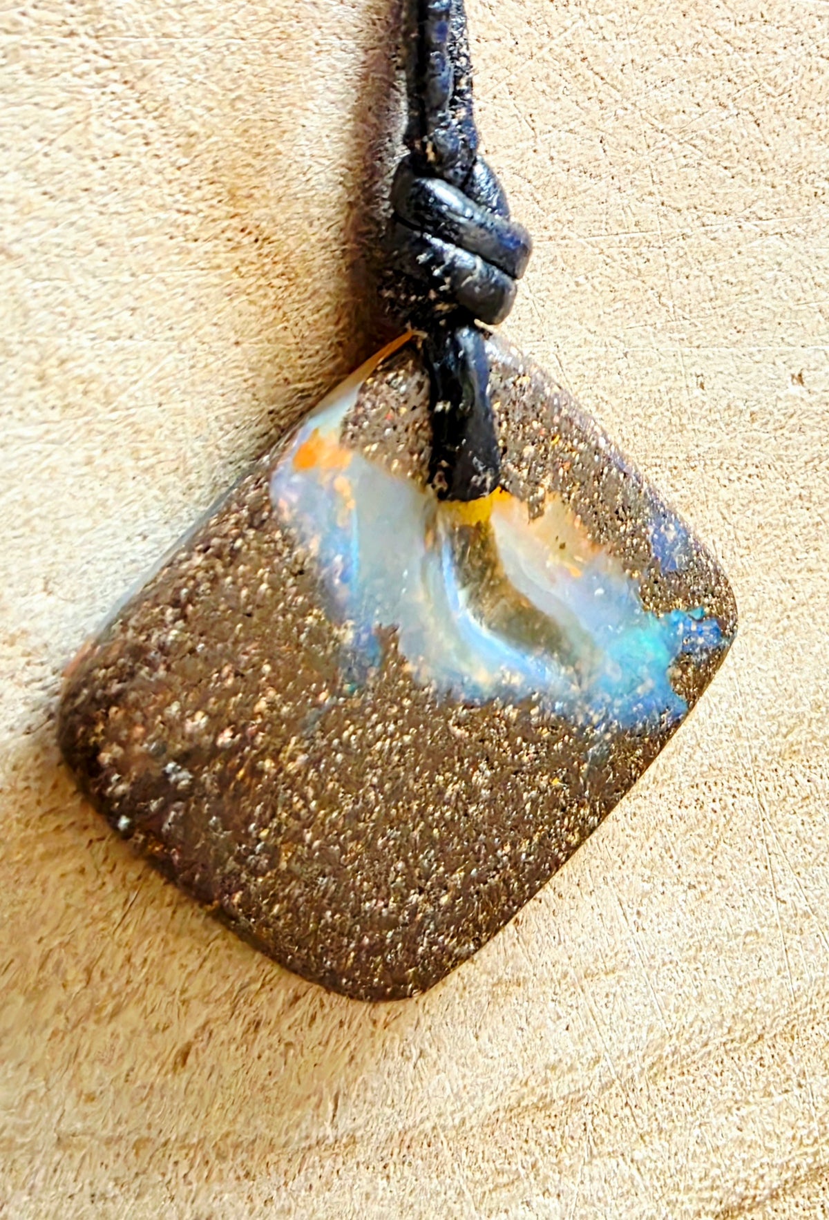A square pendant with rounded corners, featuring a textured dark brown and gold speckled pattern with a swirling iridescent blue, white, and orange inlay, suspended from a black cord on a beige textured background.