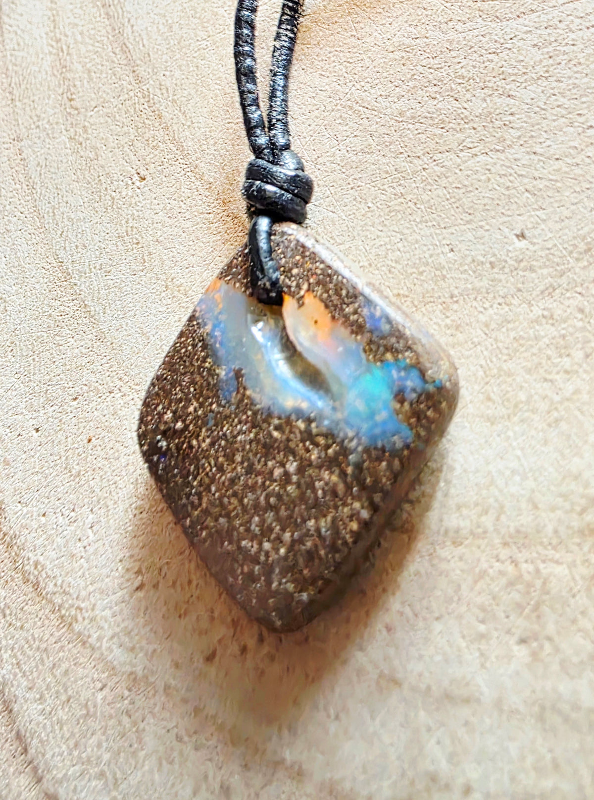 A square pendant necklace made from a textured brown stone with iridescent blue, orange, and white flashes, hanging from a black cord on a light wood background.