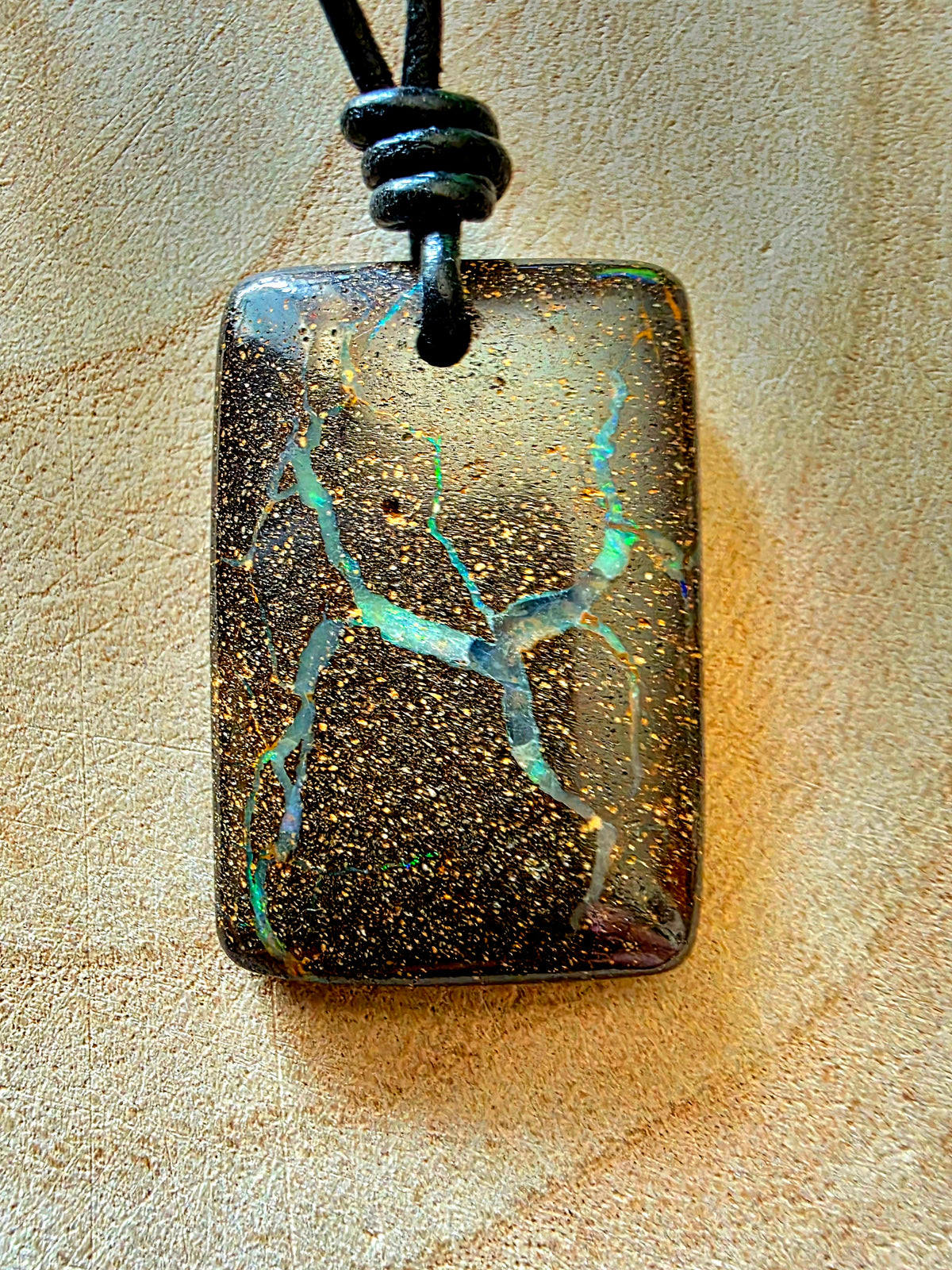 A rectangular pendant necklace featuring a dark, glittery opal stone with vibrant green and blue iridescent veins, suspended from a black cord on a textured beige background.