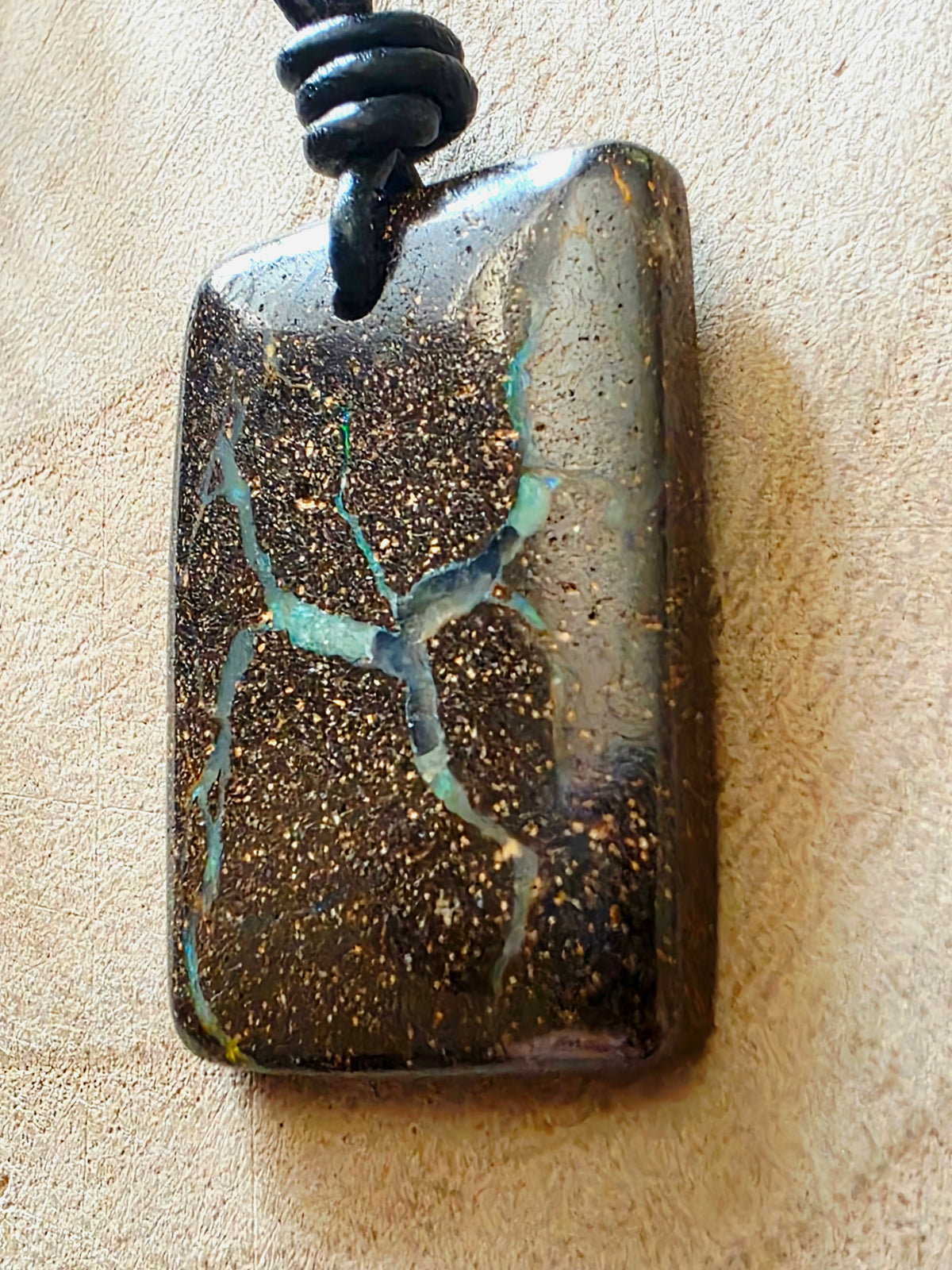 A rectangular polished stone pendant with a black cord, featuring a dark brown and black speckled body with vibrant turquoise and gold matrix veins, resting on a textured beige surface.