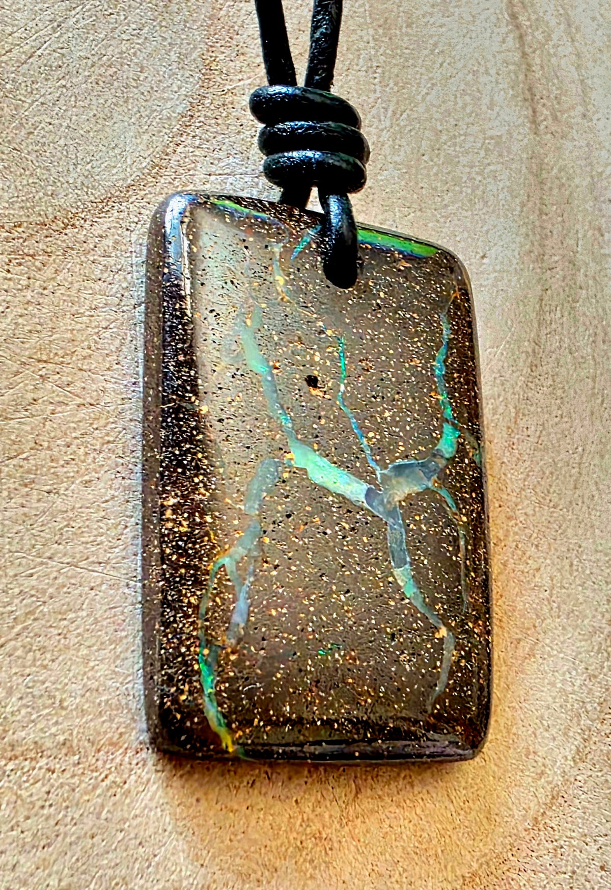 A rectangular pendant necklace featuring a dark brown, gold-speckled boulder opal with vibrant green and blue iridescent veining, suspended from a black cord on a textured beige background.