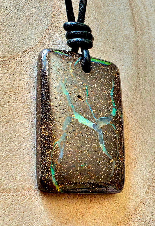 A rectangular pendant necklace featuring a dark brown, gold-speckled boulder opal with vibrant green and blue iridescent veining, suspended from a black cord on a textured beige background.