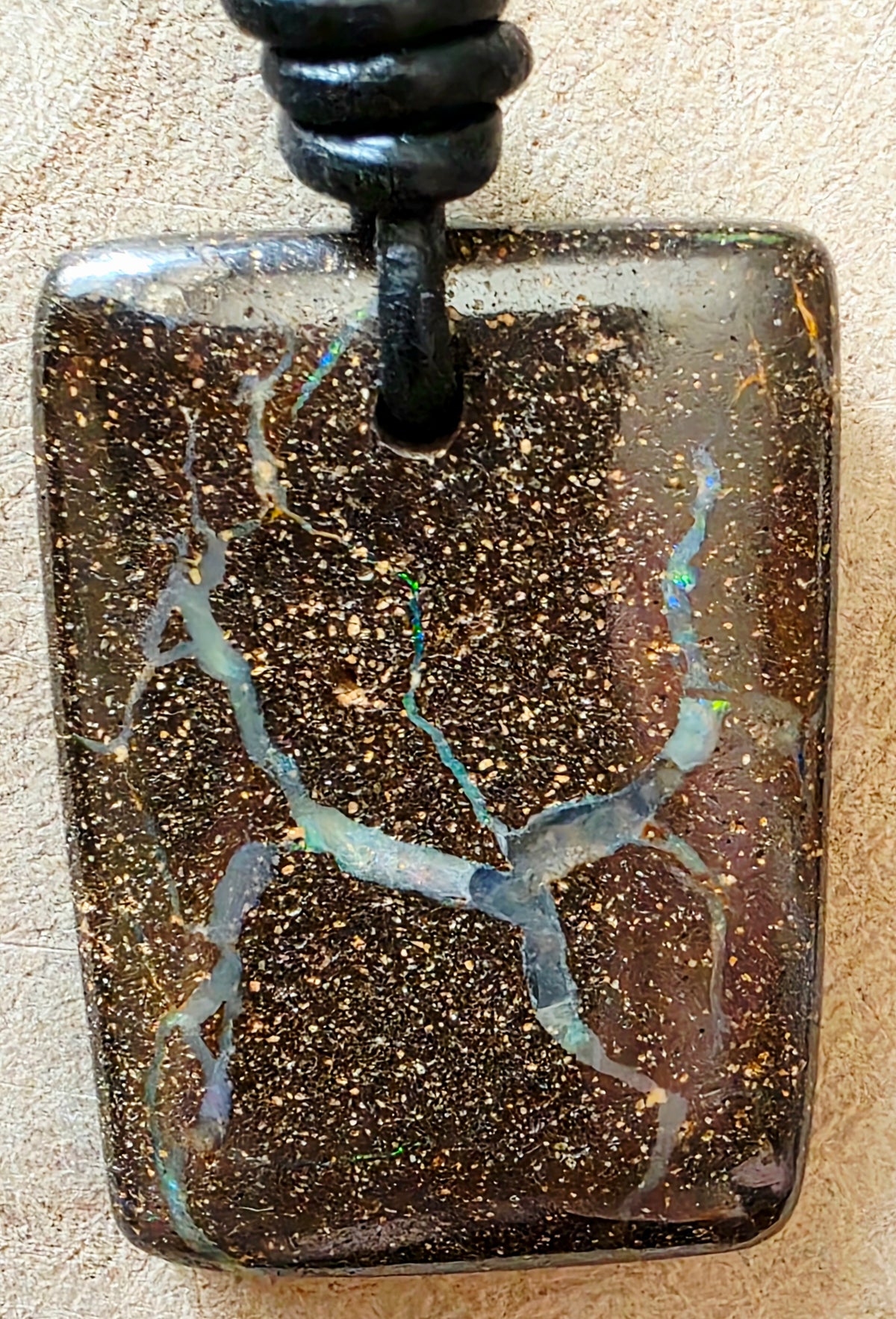 A rectangular dark brown opal pendant necklace with shimmering gold flecks and branching iridescent blue-green veining, suspended from a black cord against a light beige textured background.
