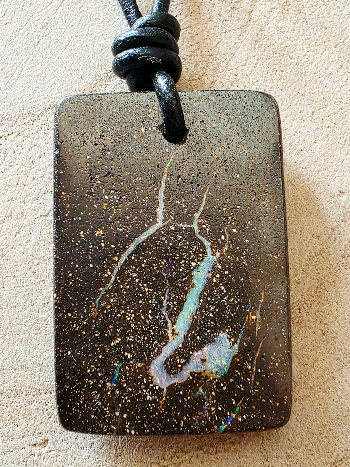 A rectangular black opal pendant with a natural gold speckled pattern and a light blue iridescent vein, hanging from a black cord on a textured beige background.