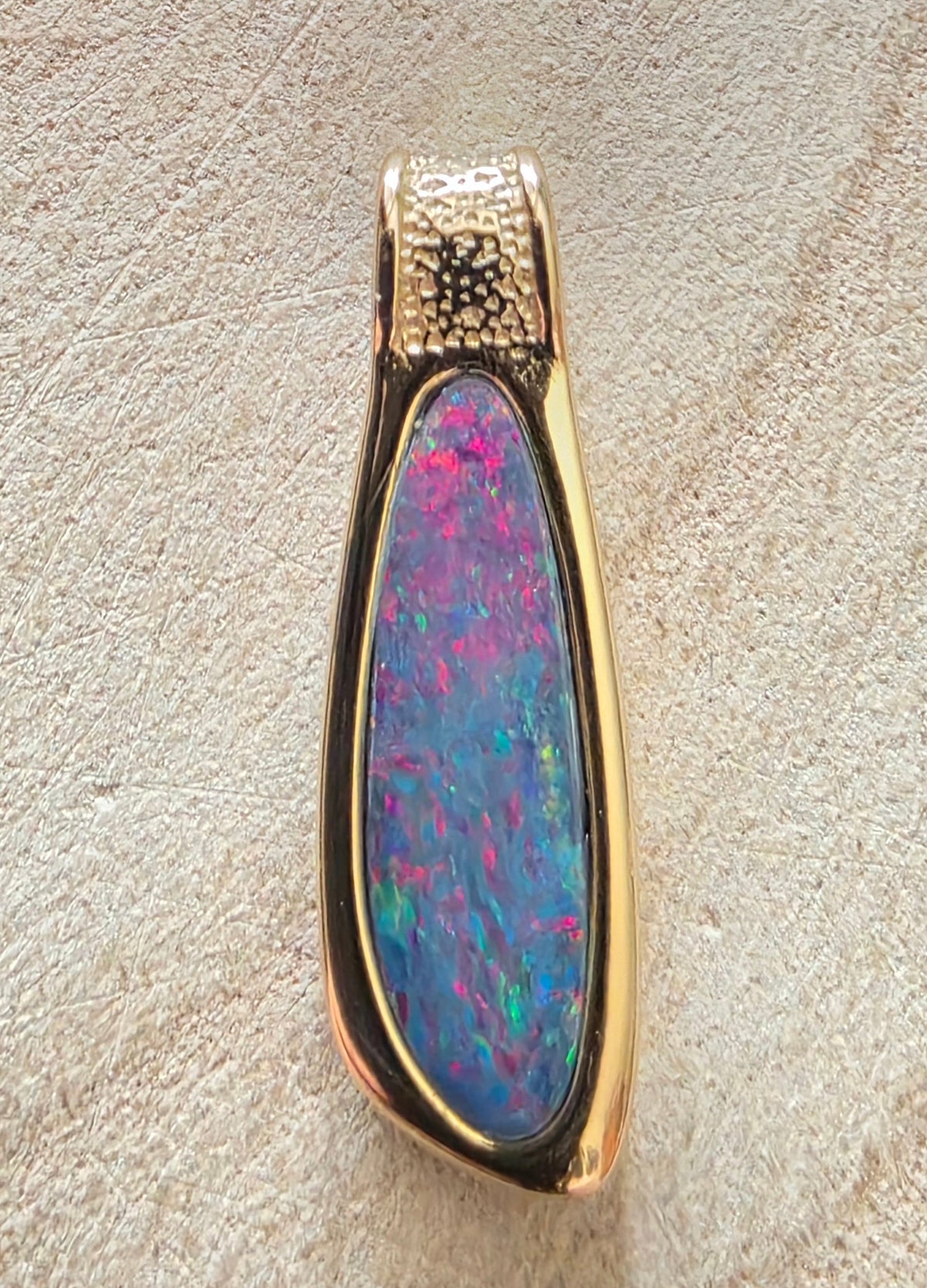 A teardrop-shaped pendant featuring a vibrant iridescent opal inlay with blue, pink, and green flashes, set in a polished gold-tone bezel with a textured gold bail, shown on a textured beige surface.