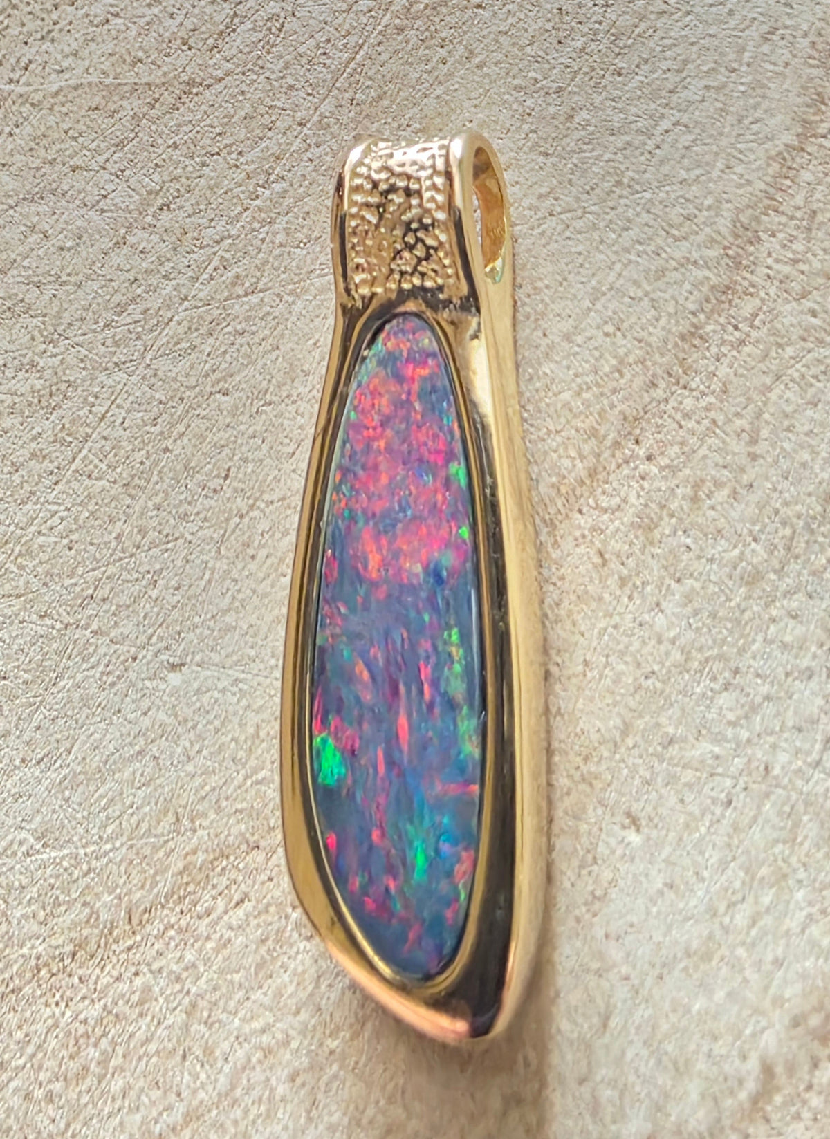 A teardrop-shaped pendant featuring a vibrant, iridescent opal stone with flashes of pink, green, and blue, set in a polished gold-tone bezel with a textured bail at the top, resting on a textured beige surface.