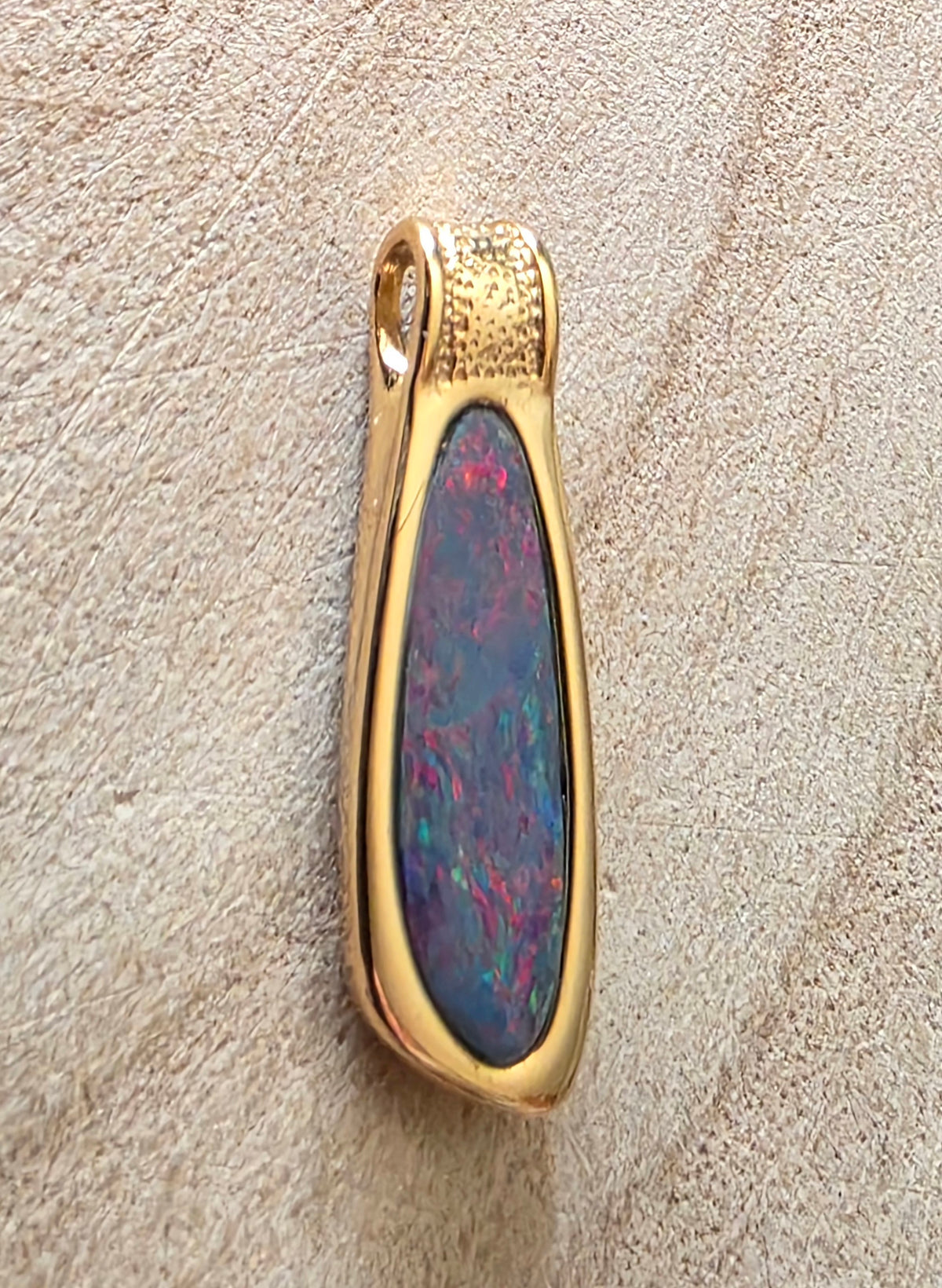 A gold pendant featuring a teardrop-shaped opal stone with vibrant blue, green, and pink flashes, set against a textured beige background.