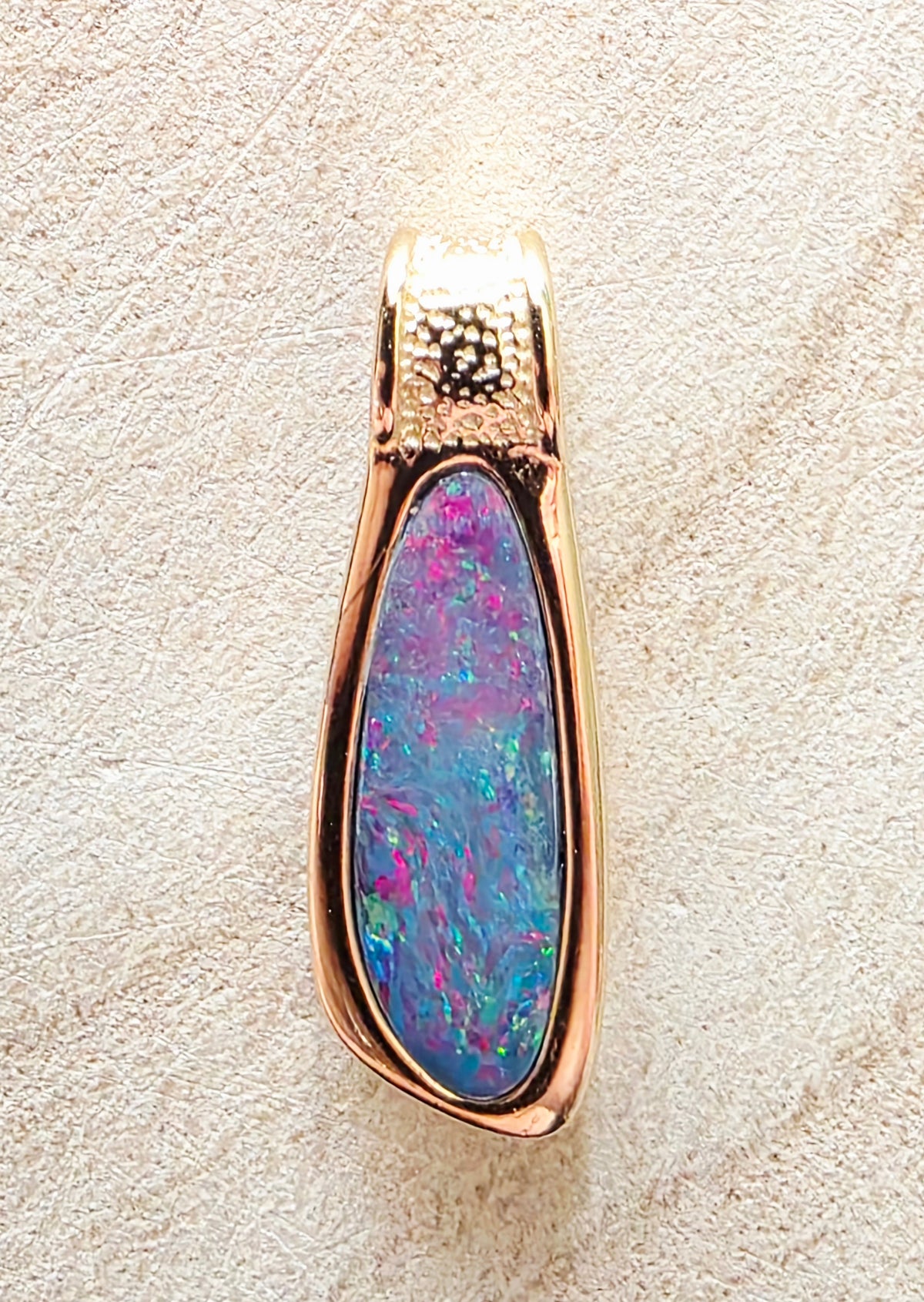 A teardrop-shaped pendant featuring a vibrant blue and purple opal inlay set in a polished rose gold-tone metal frame, displayed on a textured beige surface.