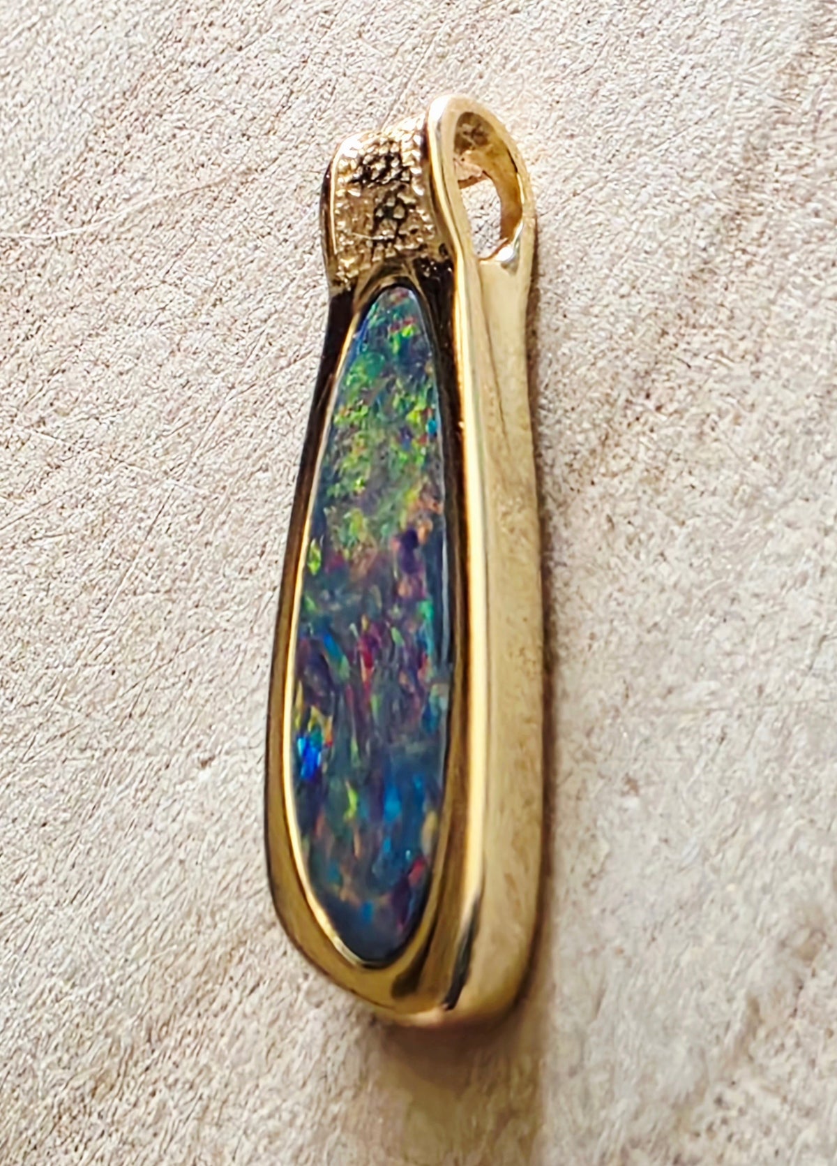 A gold pendant featuring a teardrop-shaped iridescent opal stone with vibrant blue, green, and purple flashes, set in a polished gold bezel with a textured gold top cap, resting on a textured beige surface.