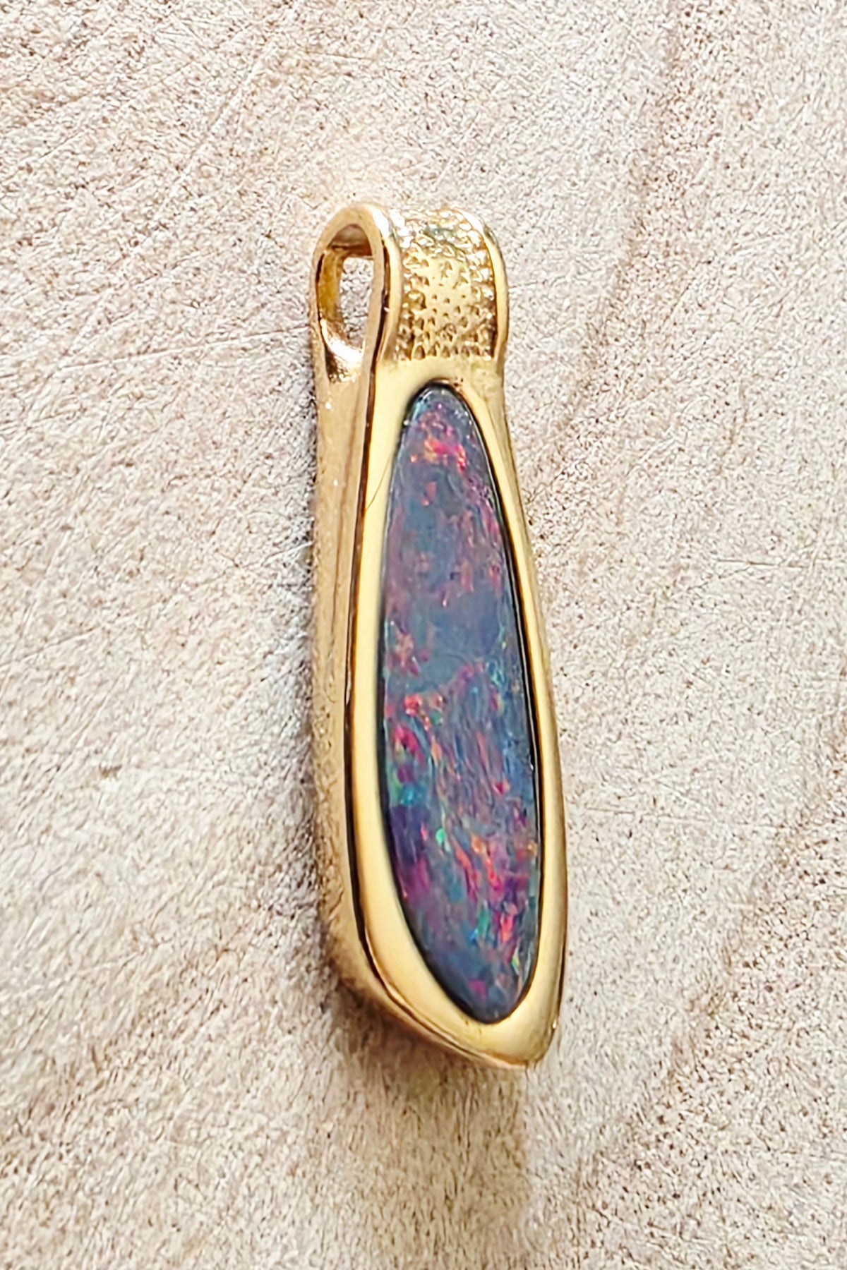 A gold pendant with a teardrop-shaped iridescent opal inlay, displayed on a textured beige surface.