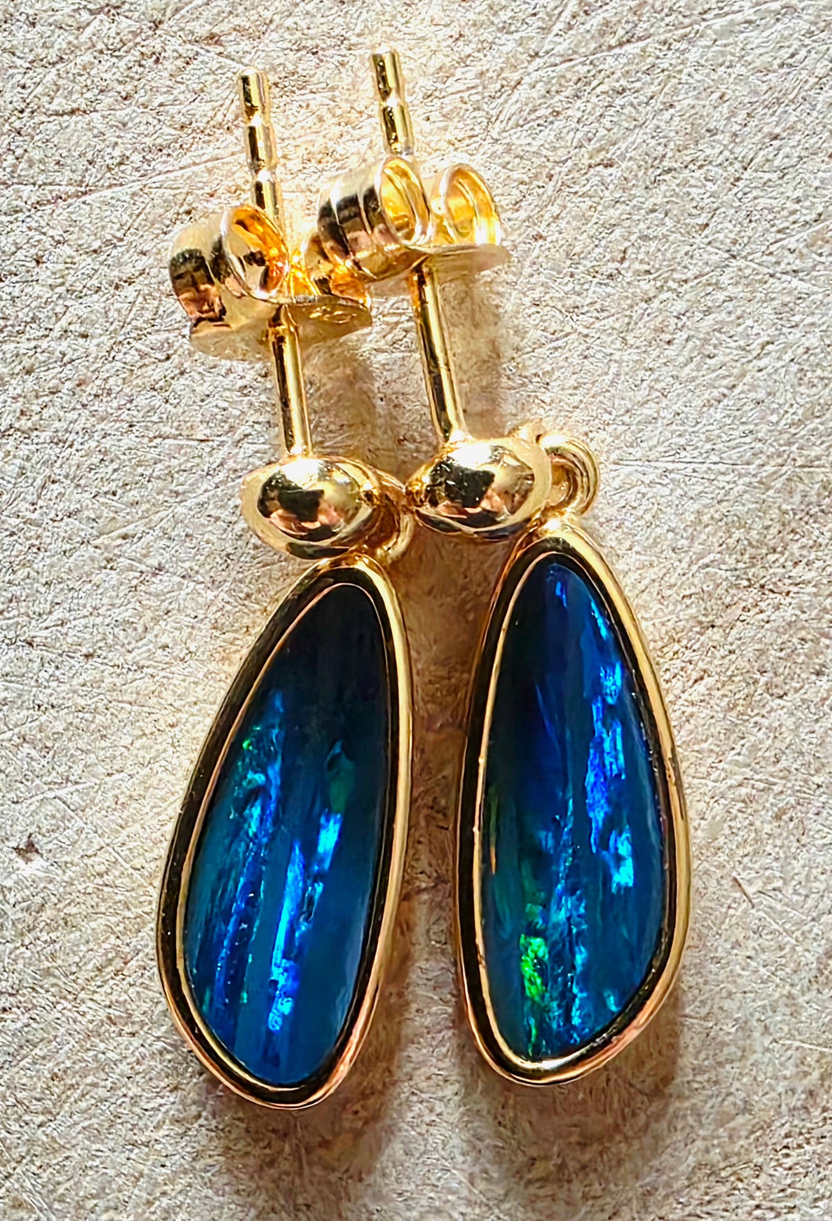 A pair of gold teardrop dangle earrings with vibrant blue iridescent opal inlays, shown on a textured beige surface.