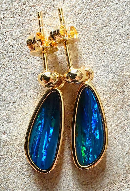 A pair of gold teardrop dangle earrings with vibrant blue iridescent opal inlays, shown on a textured beige surface.