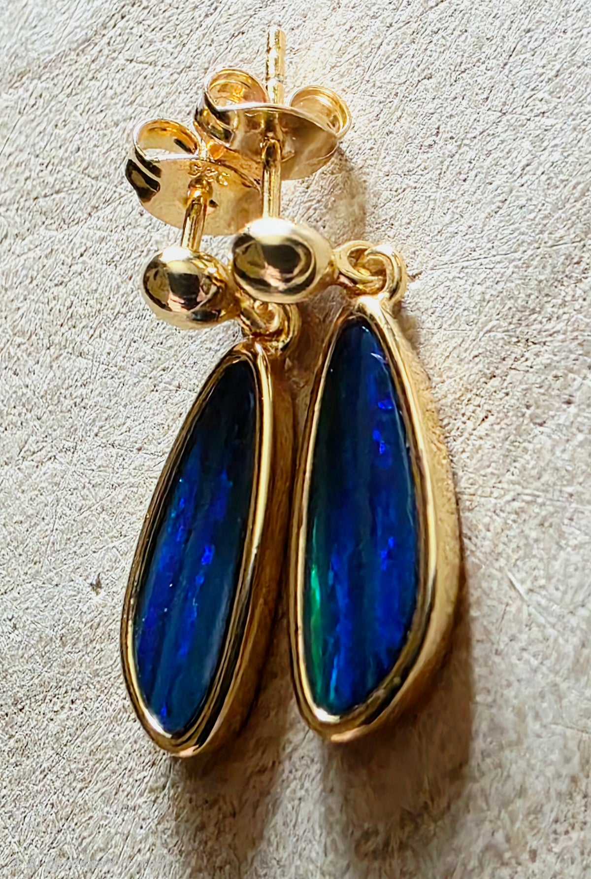 A pair of gold teardrop dangle earrings with vibrant blue iridescent opal inlays, displayed on a textured beige surface.