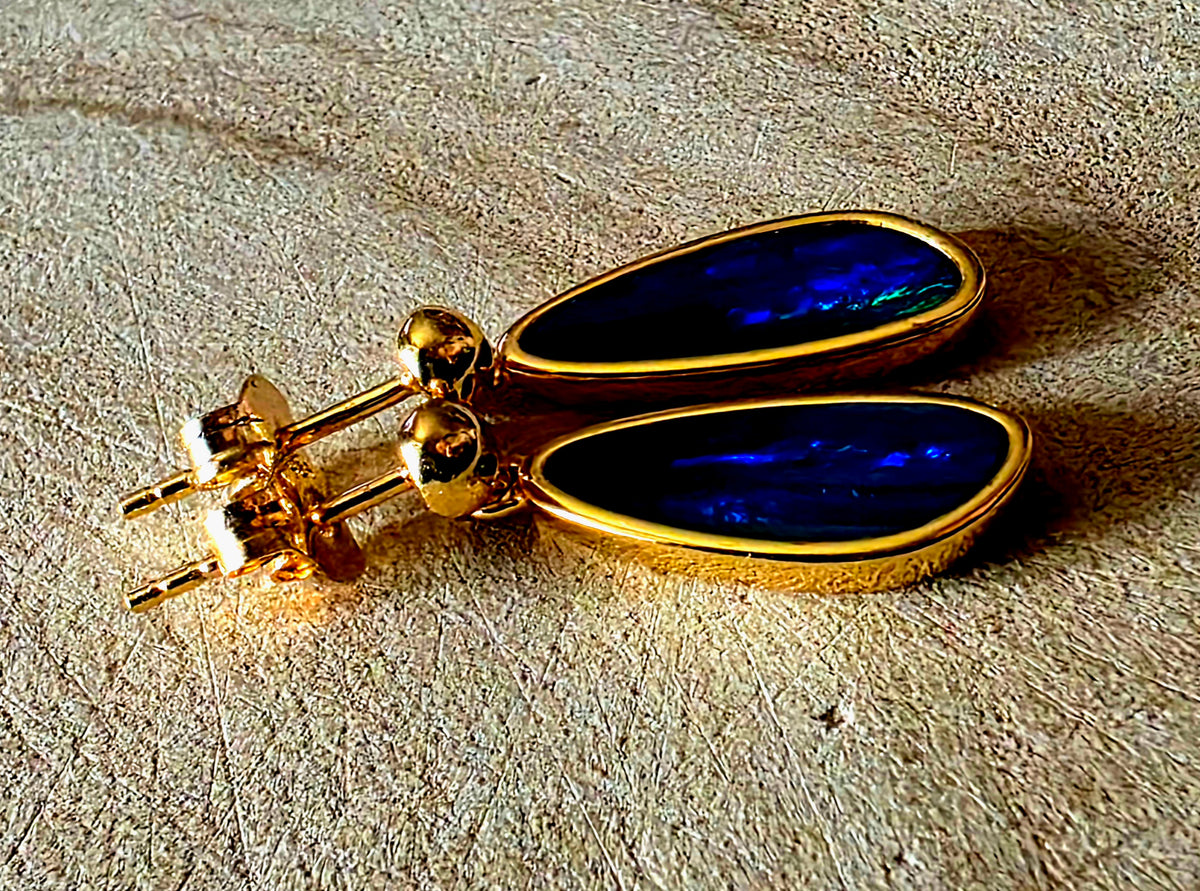 A pair of gold teardrop dangle earrings with deep blue iridescent opal inlays, displayed on a textured beige stone surface.