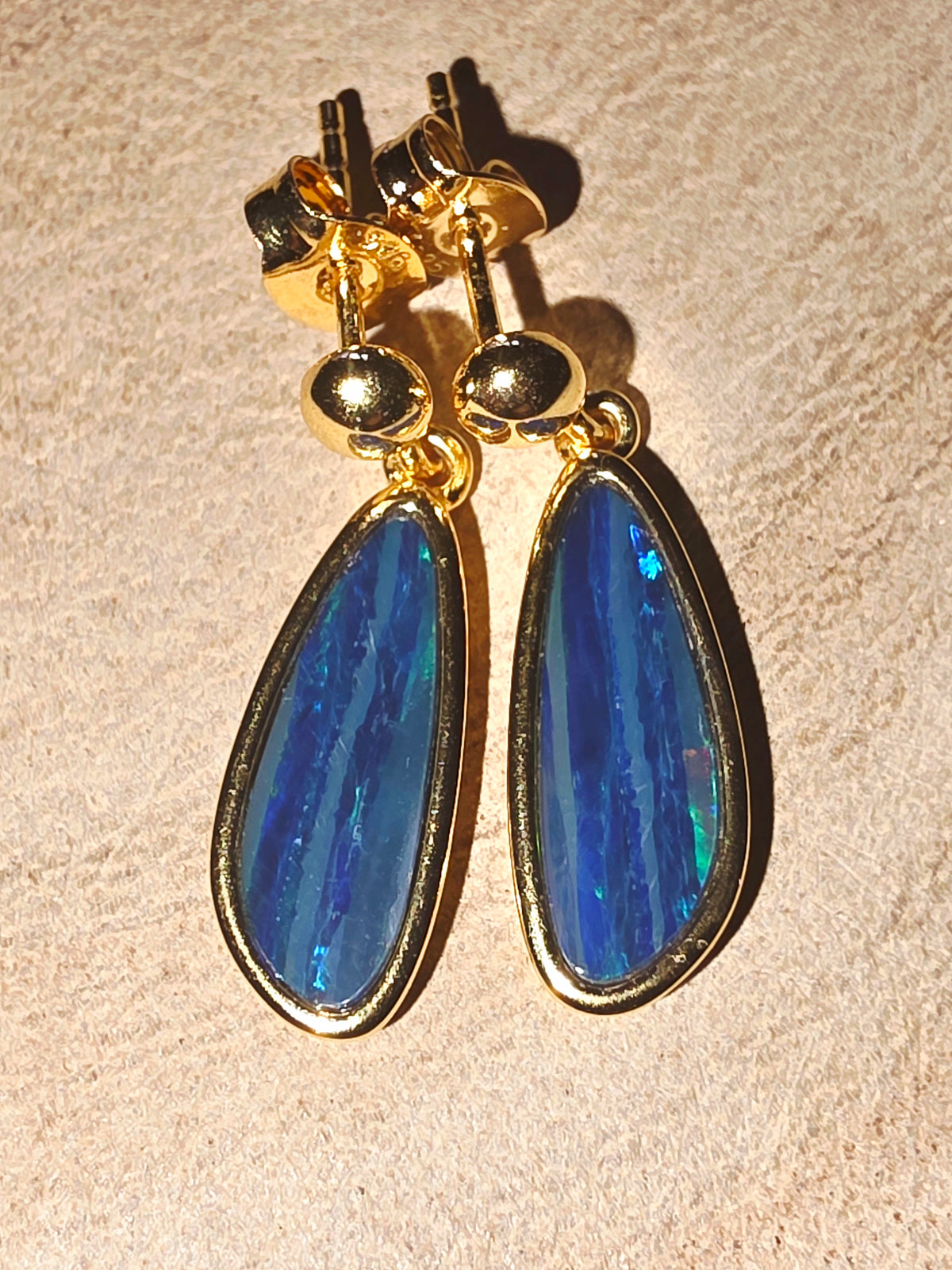 A pair of gold drop earrings with teardrop-shaped blue opal gemstones, displayed on a textured beige surface.