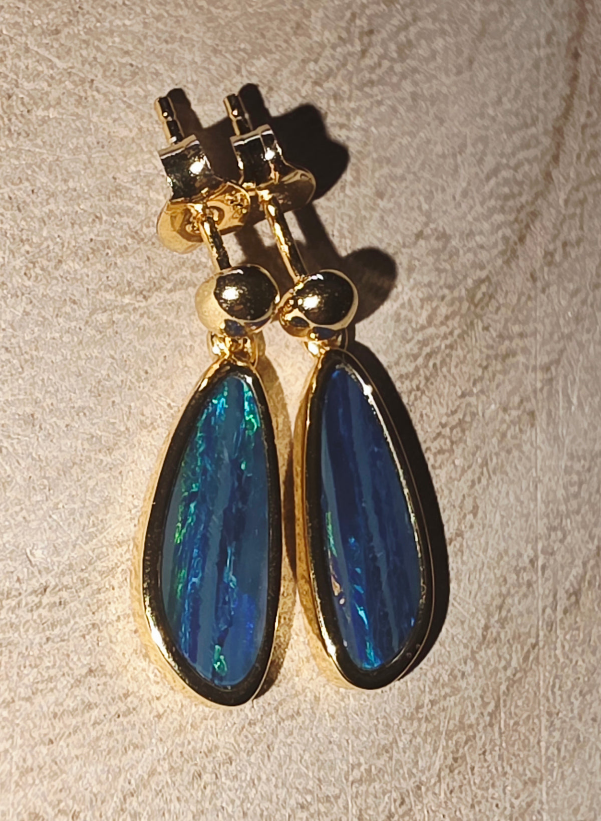A pair of gold drop earrings with teardrop-shaped blue iridescent opal stones, displayed on a textured beige surface.