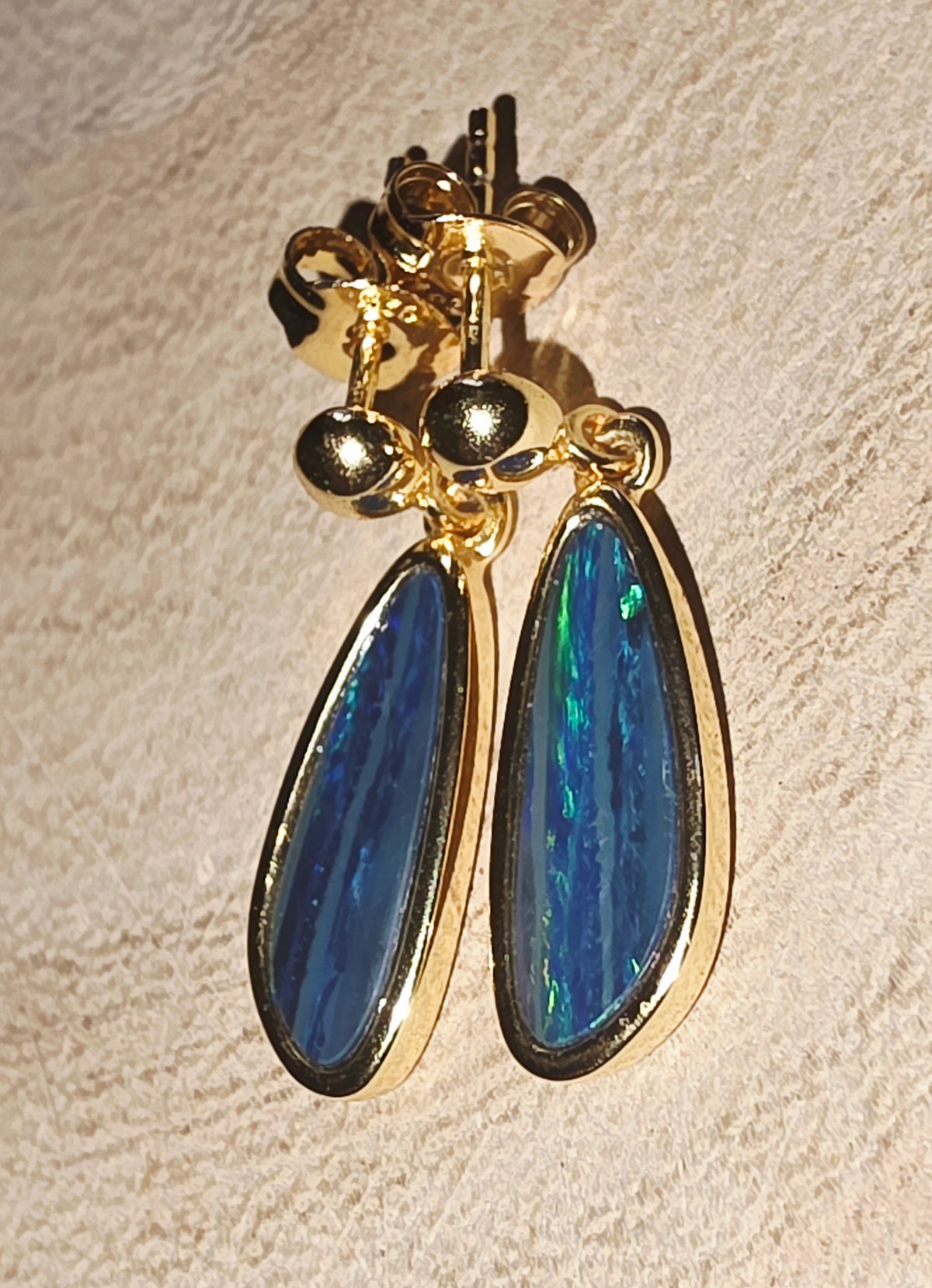 A pair of gold drop earrings with teardrop-shaped blue iridescent opal stones and small gold bead accents, shown on a textured beige surface.