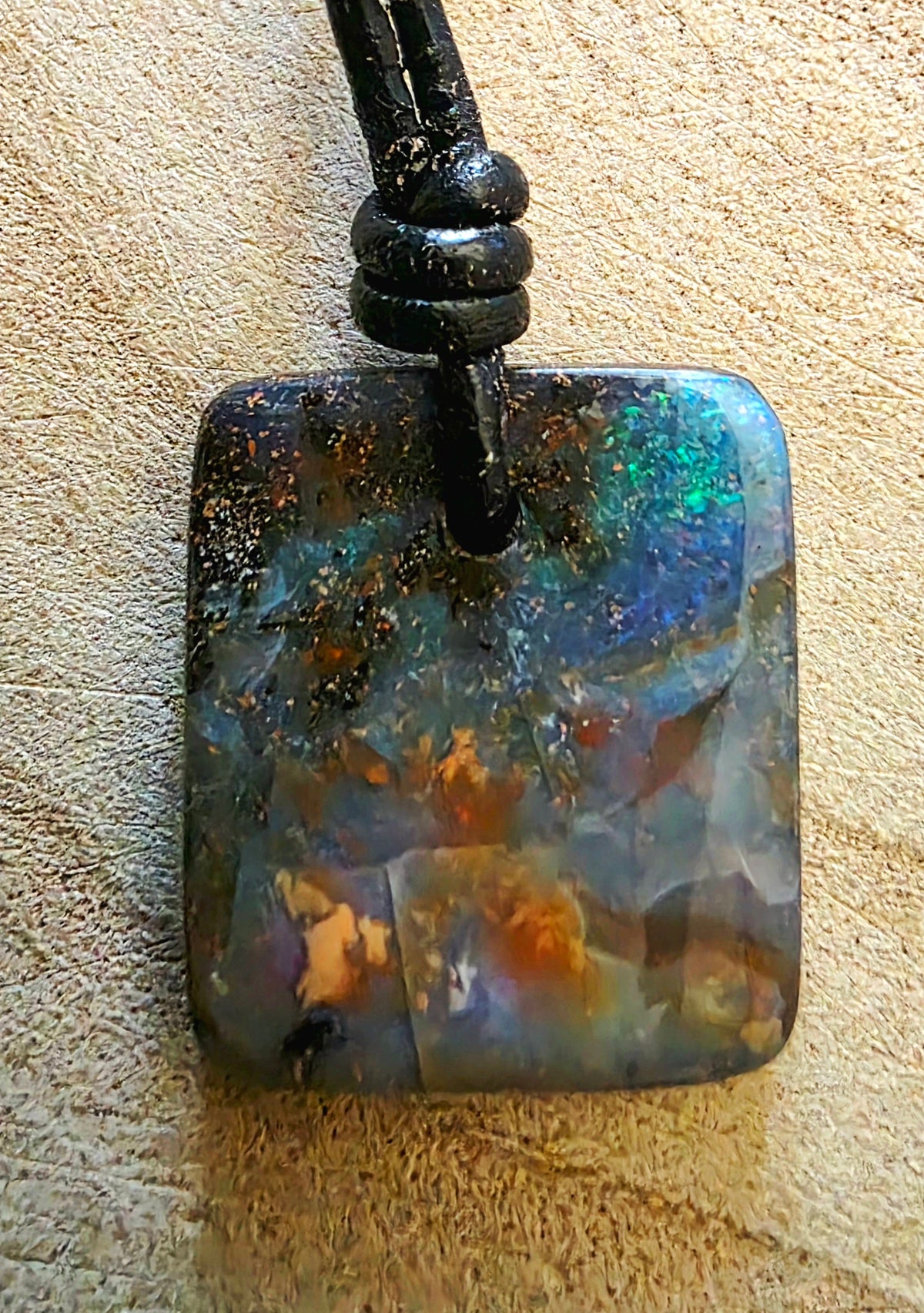 A square iridescent opal pendant necklace on a black cord, displayed on a textured beige surface.