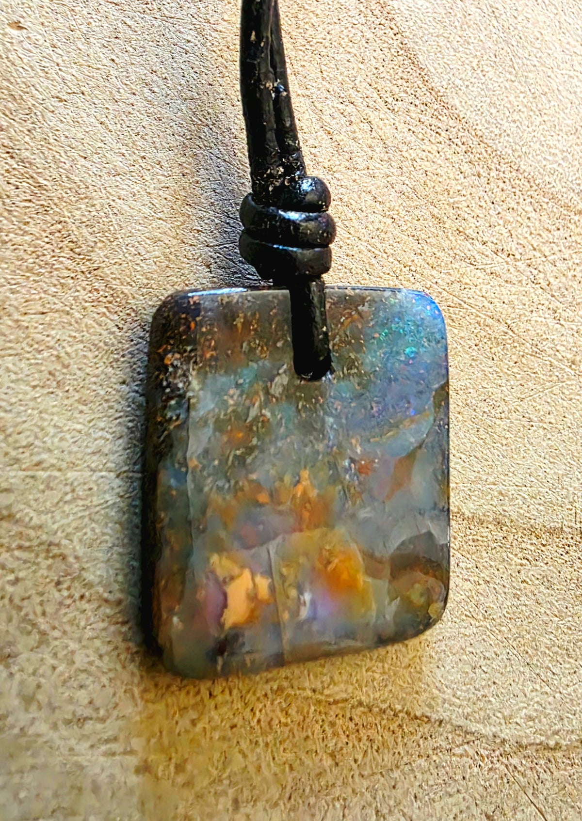 A square iridescent opal pendant with a black cord, resting on a textured beige surface.