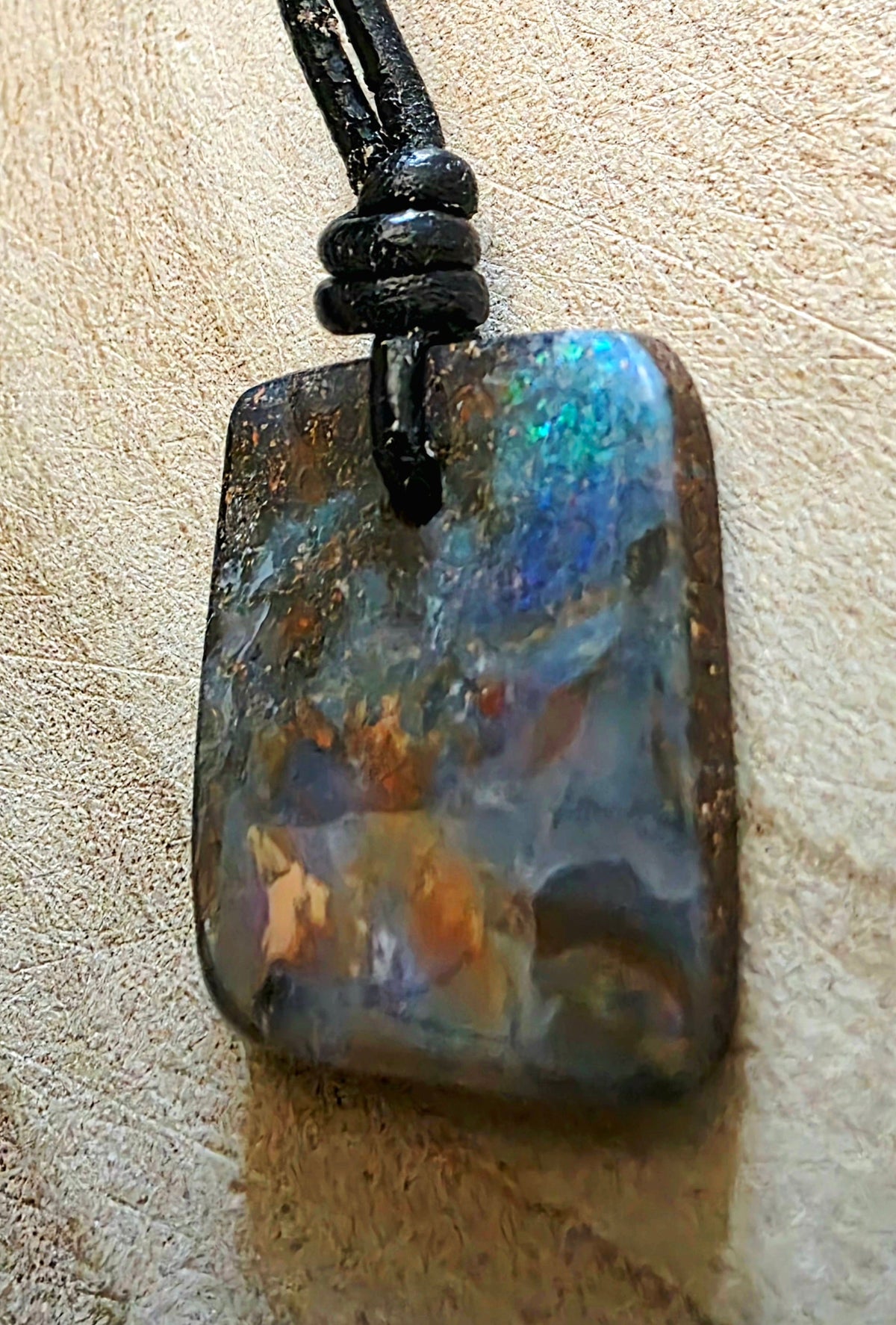 A rectangular iridescent opal pendant necklace with a black cord, resting on a textured beige surface.