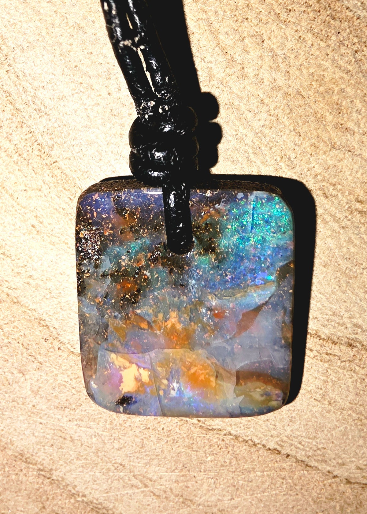 A square iridescent opal pendant necklace with a black cord, resting on a textured sandy beige background.