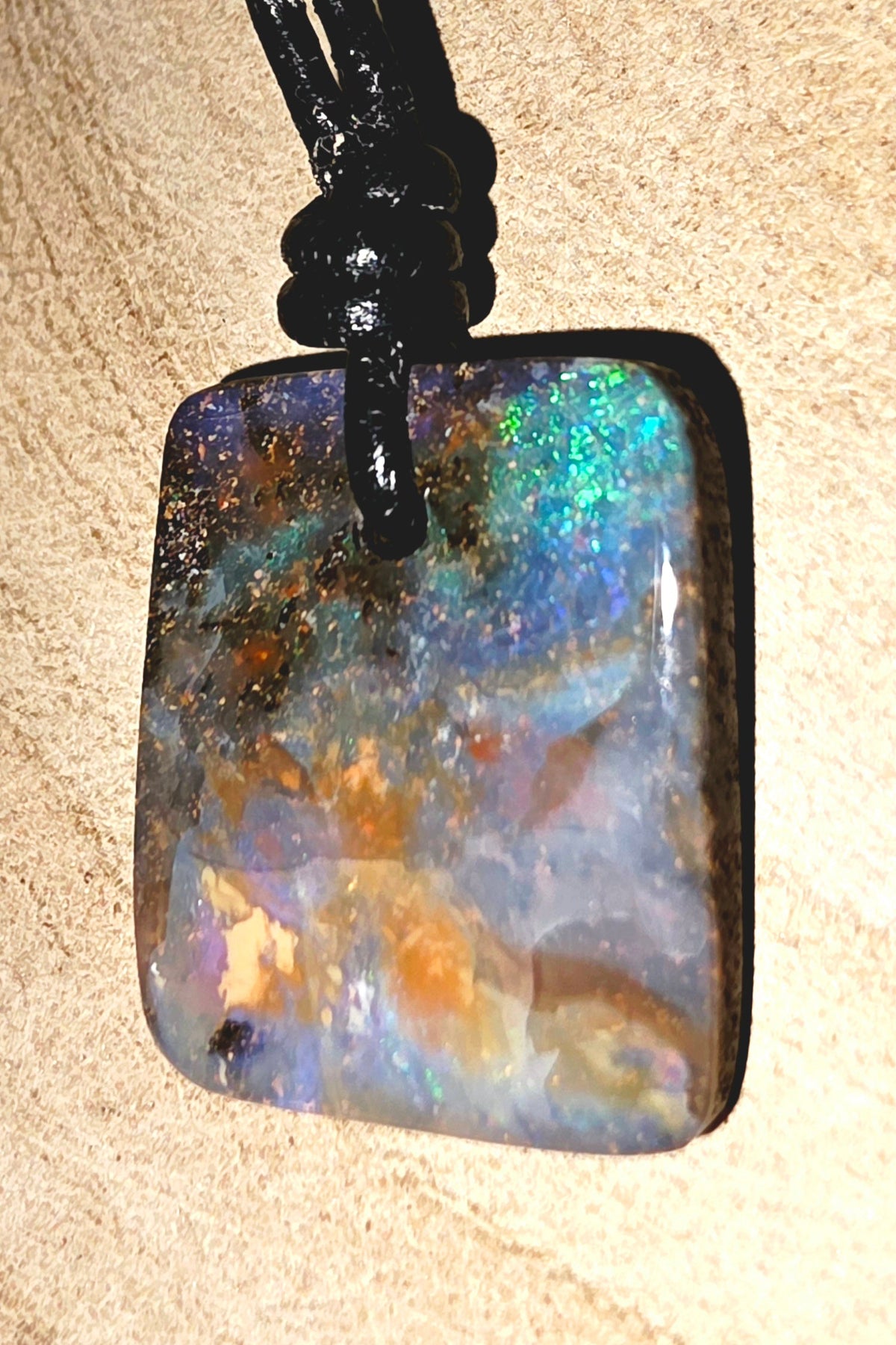 A square iridescent opal pendant necklace with a black cord, resting on a textured beige surface.