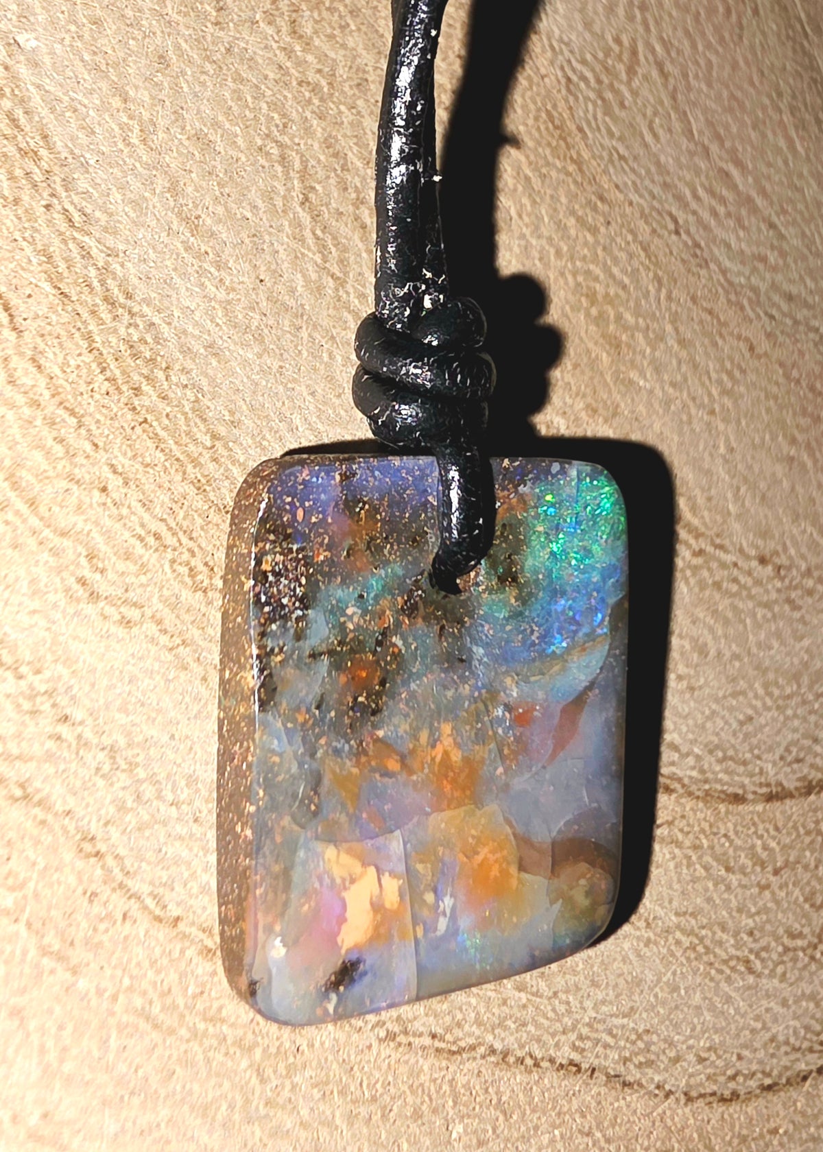 A rectangular iridescent opal pendant with a black cord, resting on a textured beige surface.