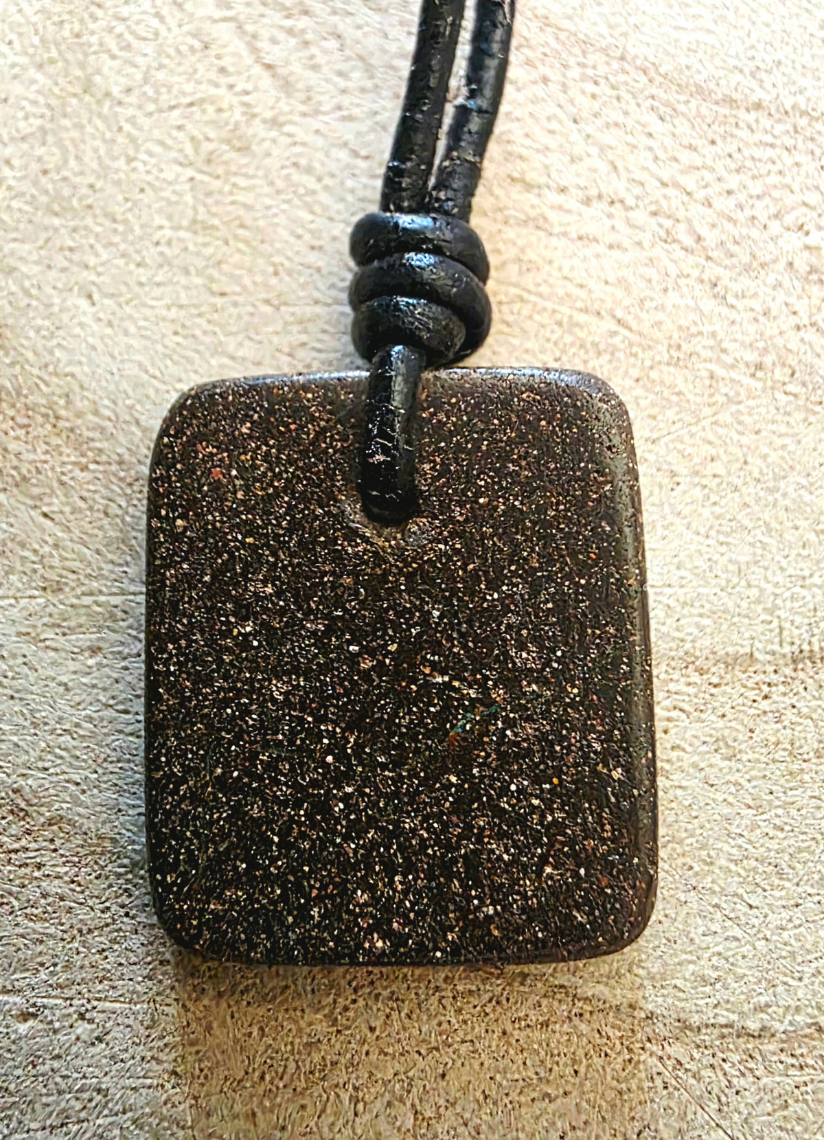 A square dark brown stone pendant with fine gold and silver flecks, suspended from a black cord, set against a textured beige background.