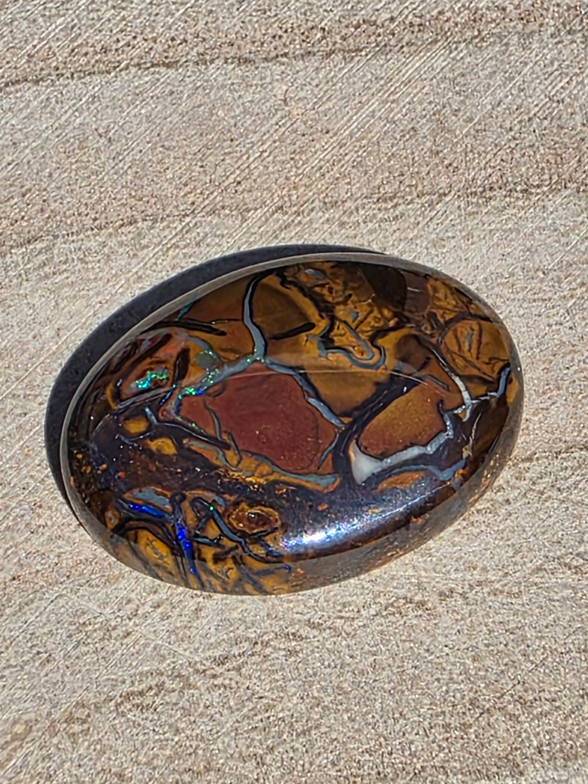 A polished oval cabochon gemstone featuring a complex pattern of metallic gold, copper, brown, and iridescent blue-green flashes, resting on a textured beige sandy surface.