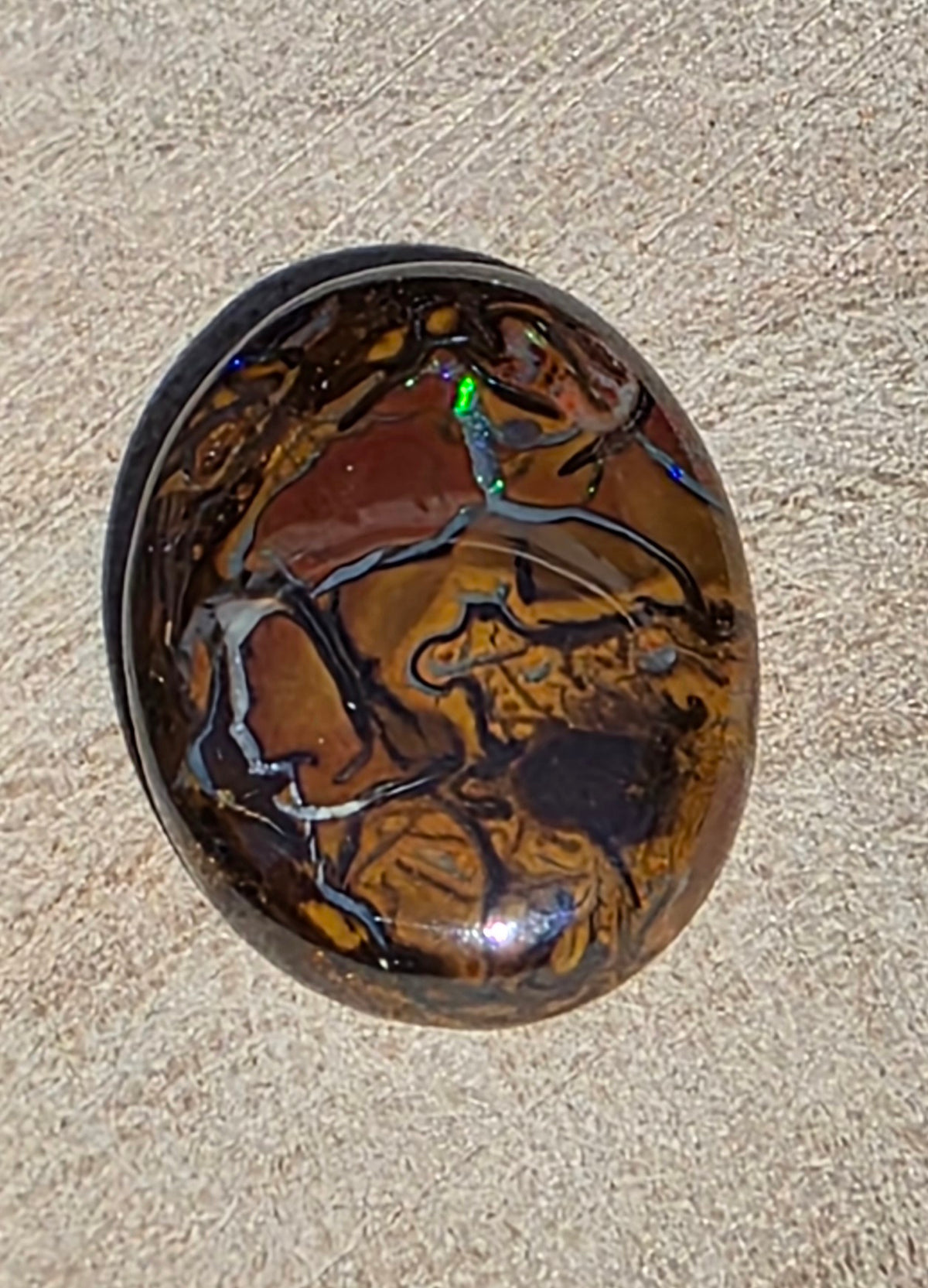A polished oval cabochon gemstone featuring a marbled pattern of rich brown, amber, and black, with a small iridescent green flash, displayed on a textured beige surface.