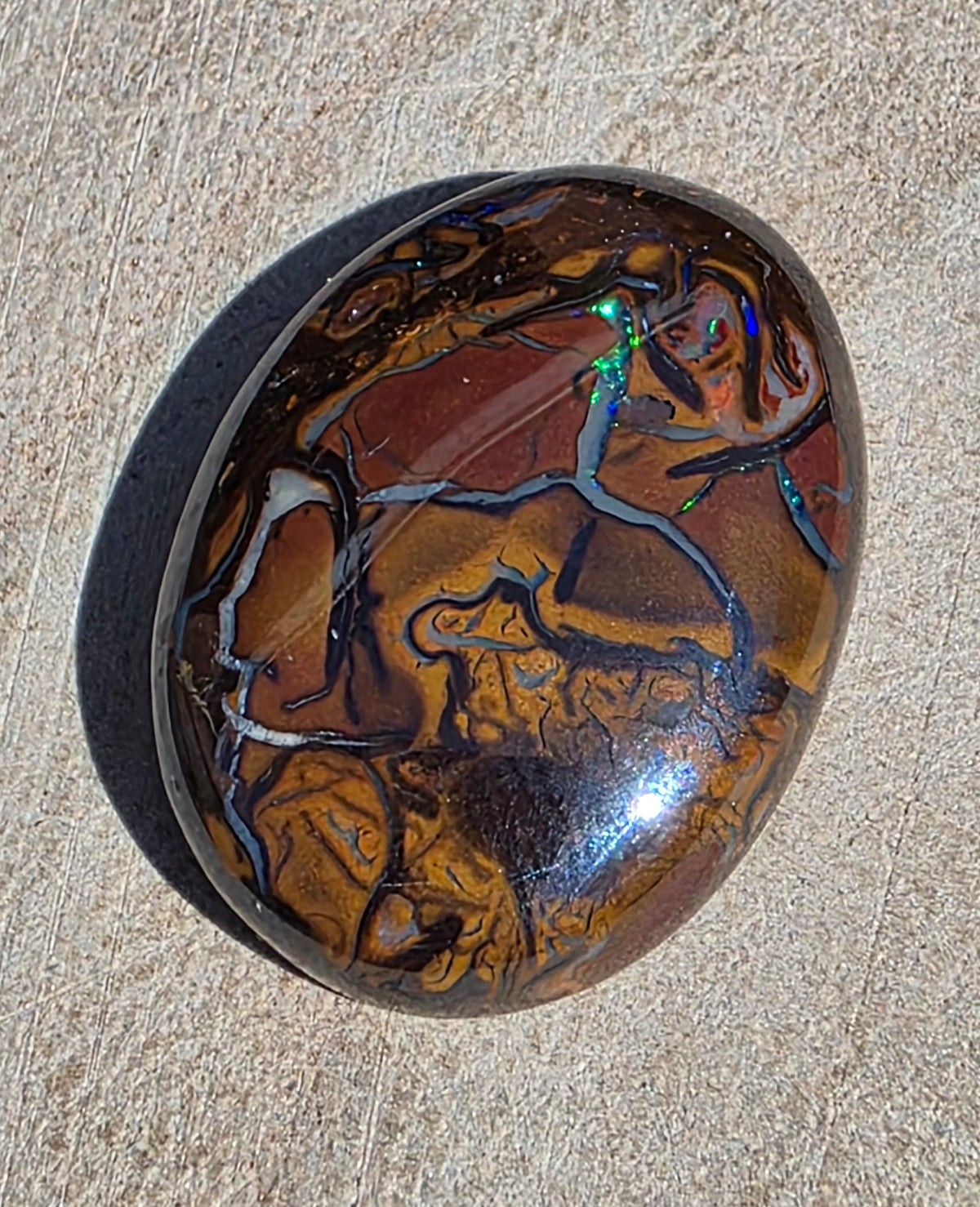 A polished oval cabochon gemstone featuring a marbled pattern of brown, amber, and black, with a vibrant iridescent play-of-colour effect, resting on a textured beige stone surface.