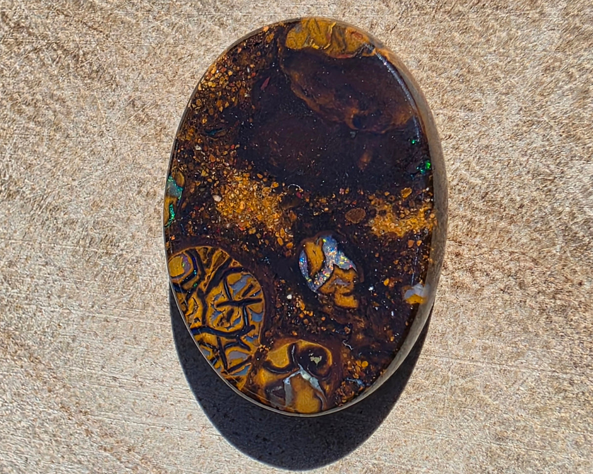 An oval cabochon gemstone featuring a marbled pattern of dark brown, black, and vibrant golden-orange, resting on a textured sandy beige surface.