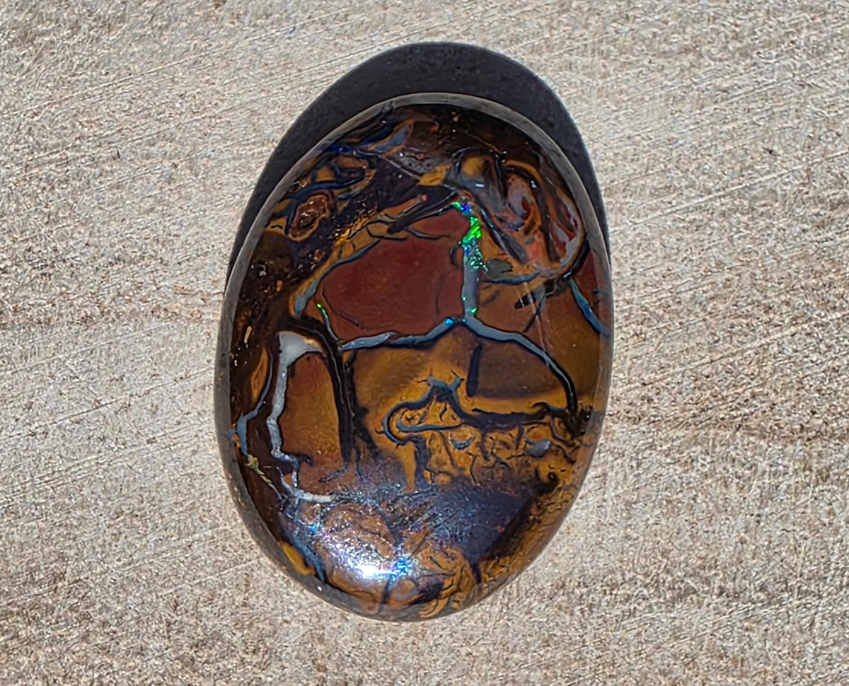 A polished oval cabochon gemstone featuring a marbled pattern of metallic gold, copper, and deep brown, with vibrant iridescent flashes of blue, green, and purple. The stone is photographed from directly above on a textured beige stone surface.