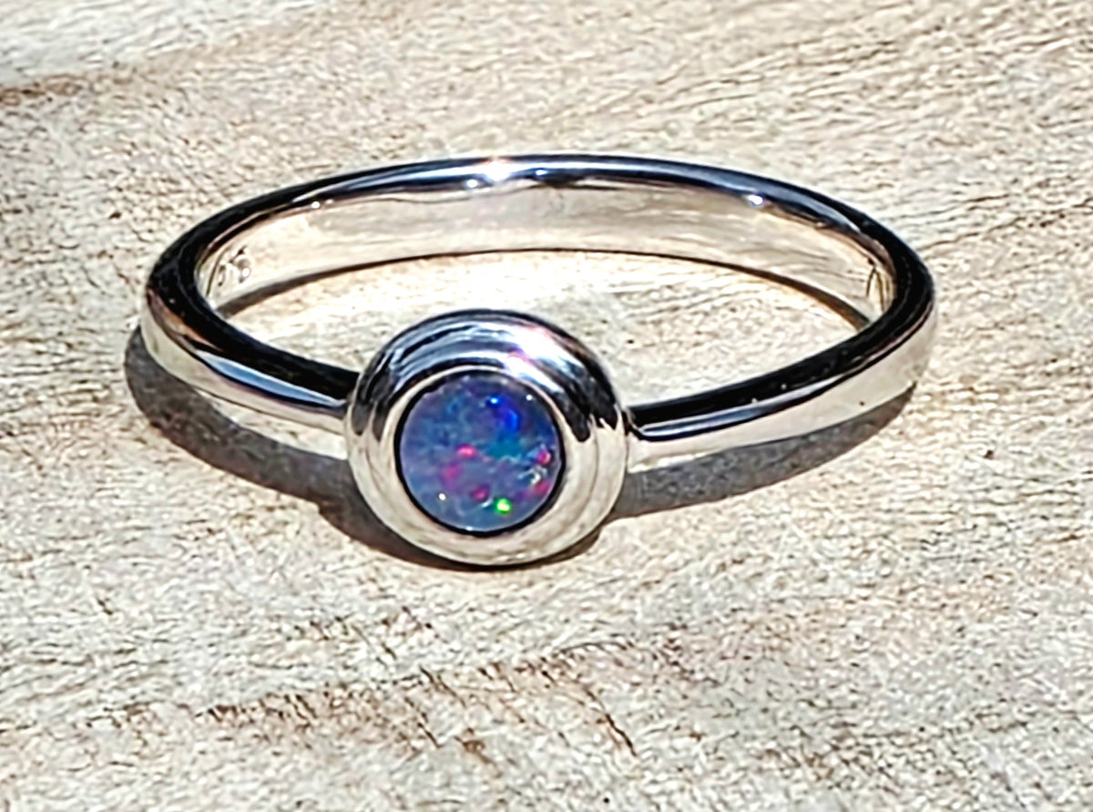 A polished sterling silver ring featuring a bezel-set round cabochon opal with vibrant blue, green, and pink flashes, resting on a textured light beige wood surface.
