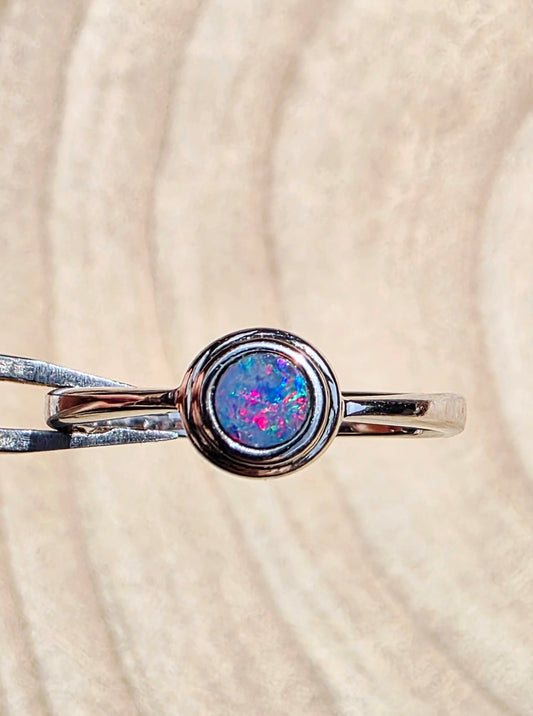 A silver ring with a round, bezel-set iridescent opal gemstone held by metal tweezers against a textured beige background.