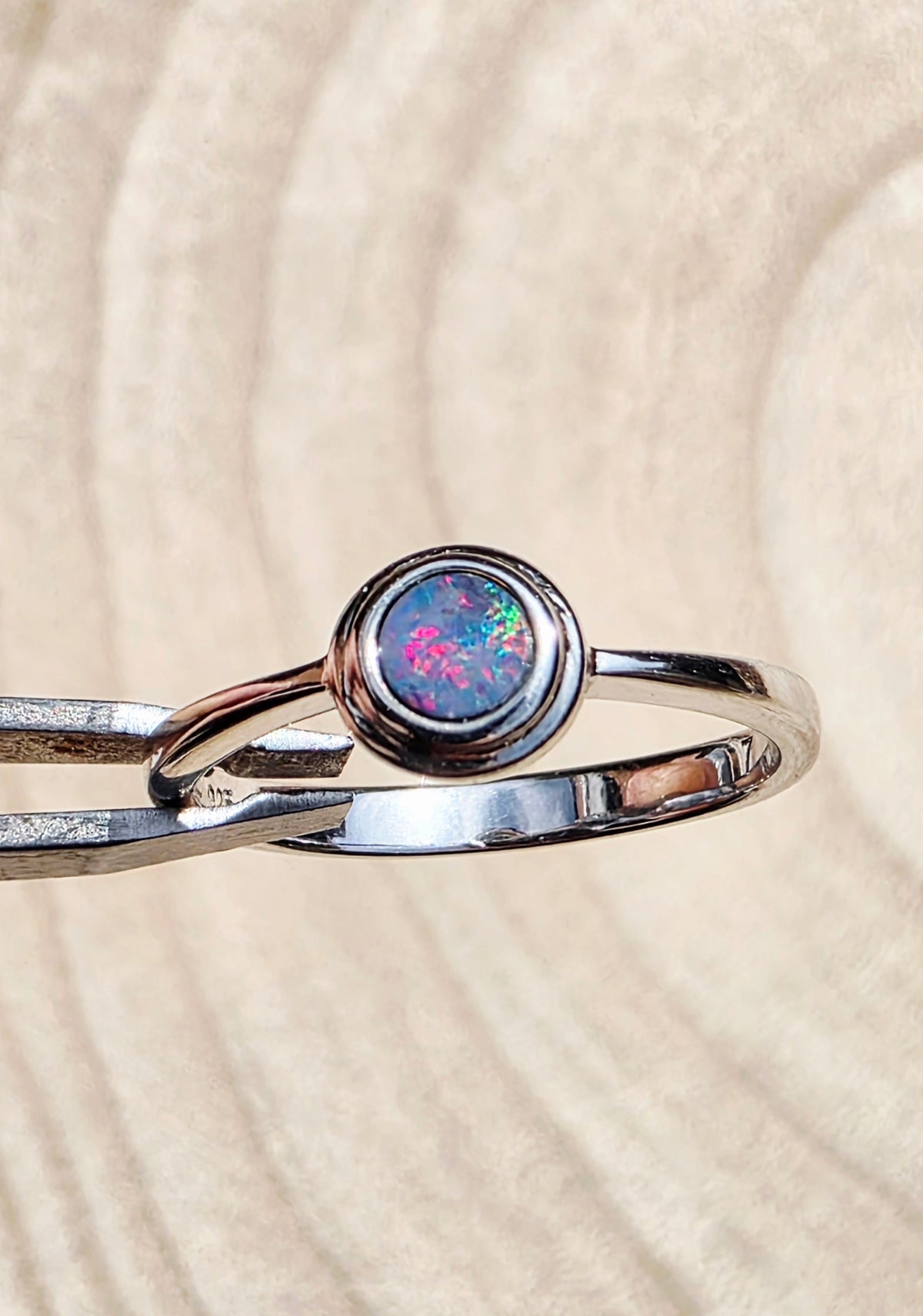 A silver ring featuring a round bezel-set iridescent opal stone with pink, blue, and green flashes, resting on a textured beige background.