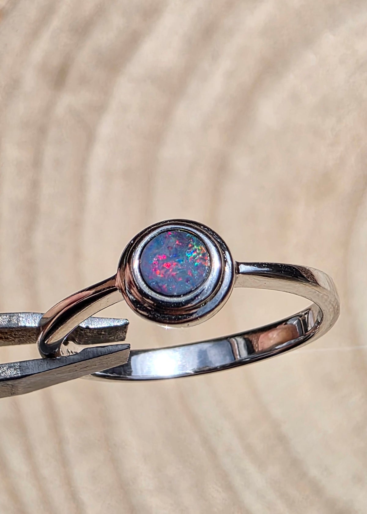 A polished sterling silver ring with a round bezel-set iridescent opal gemstone, held in forceps against a textured beige background.