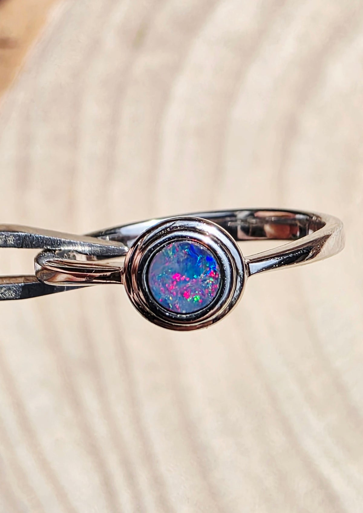 A silver ring featuring a round bezel-set iridescent opal stone with vibrant pink, blue, and green flashes, resting on a light beige wood-grain surface.