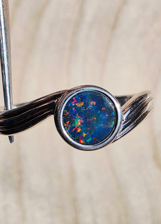 A silver ring featuring a round cabochon opal gemstone with vibrant blue, orange, and green flashes, held by a metal stand against a light wood background.