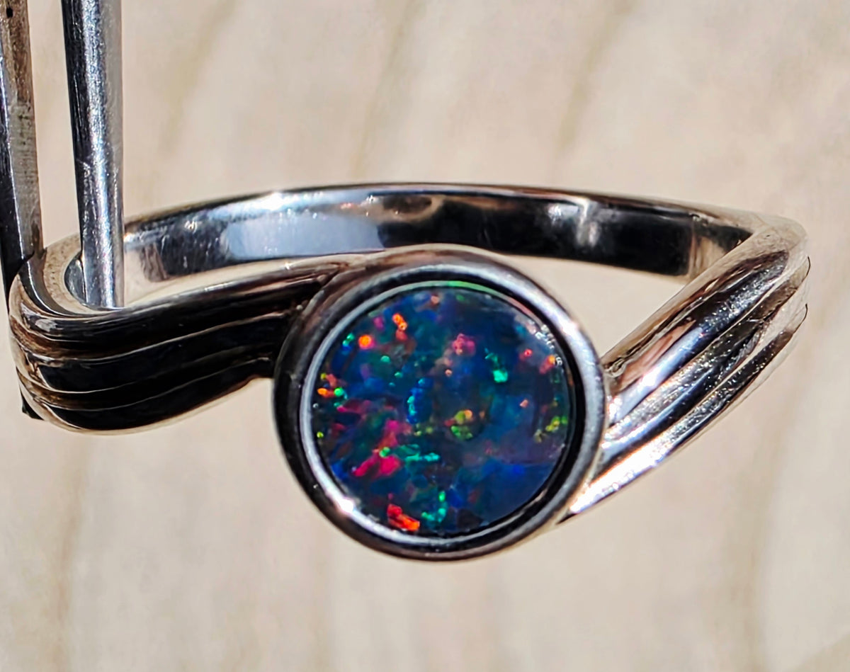 A silver ring featuring a round, vibrant iridescent opal gemstone with blue, green, red, and orange flashes, set in a polished silver band with a twisted, layered design.