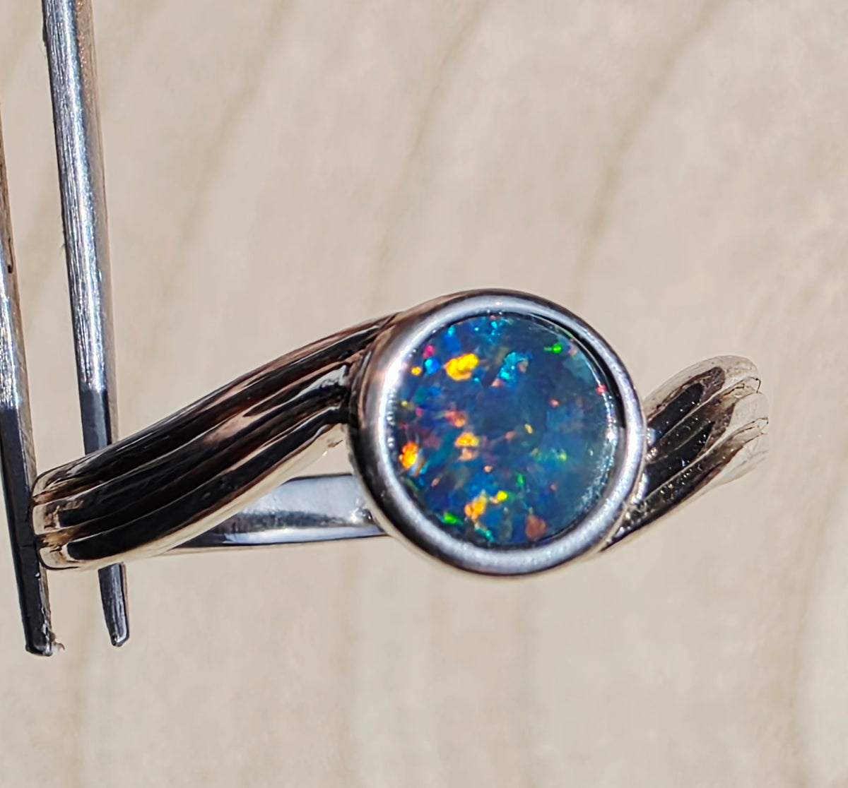 A silver ring with a round iridescent opal gemstone, held in forceps against a light beige textured background.