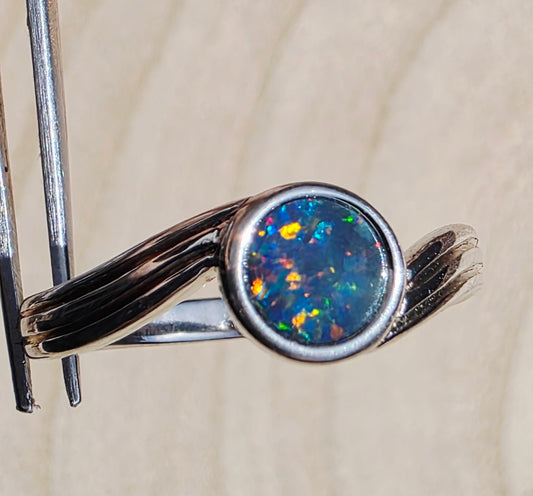A silver ring with a round iridescent opal gemstone, held in forceps against a light beige textured background.