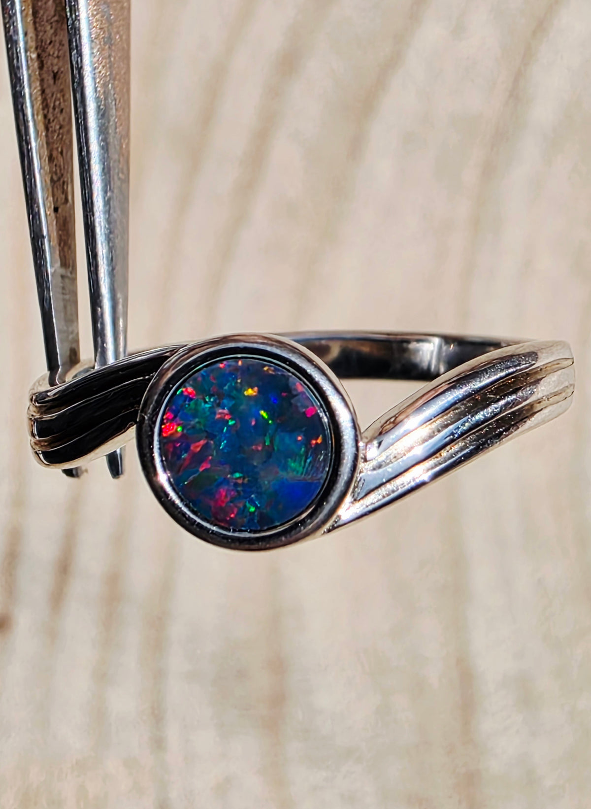A silver ring with a round iridescent opal center, held by metal tweezers against a light wood grain background.
