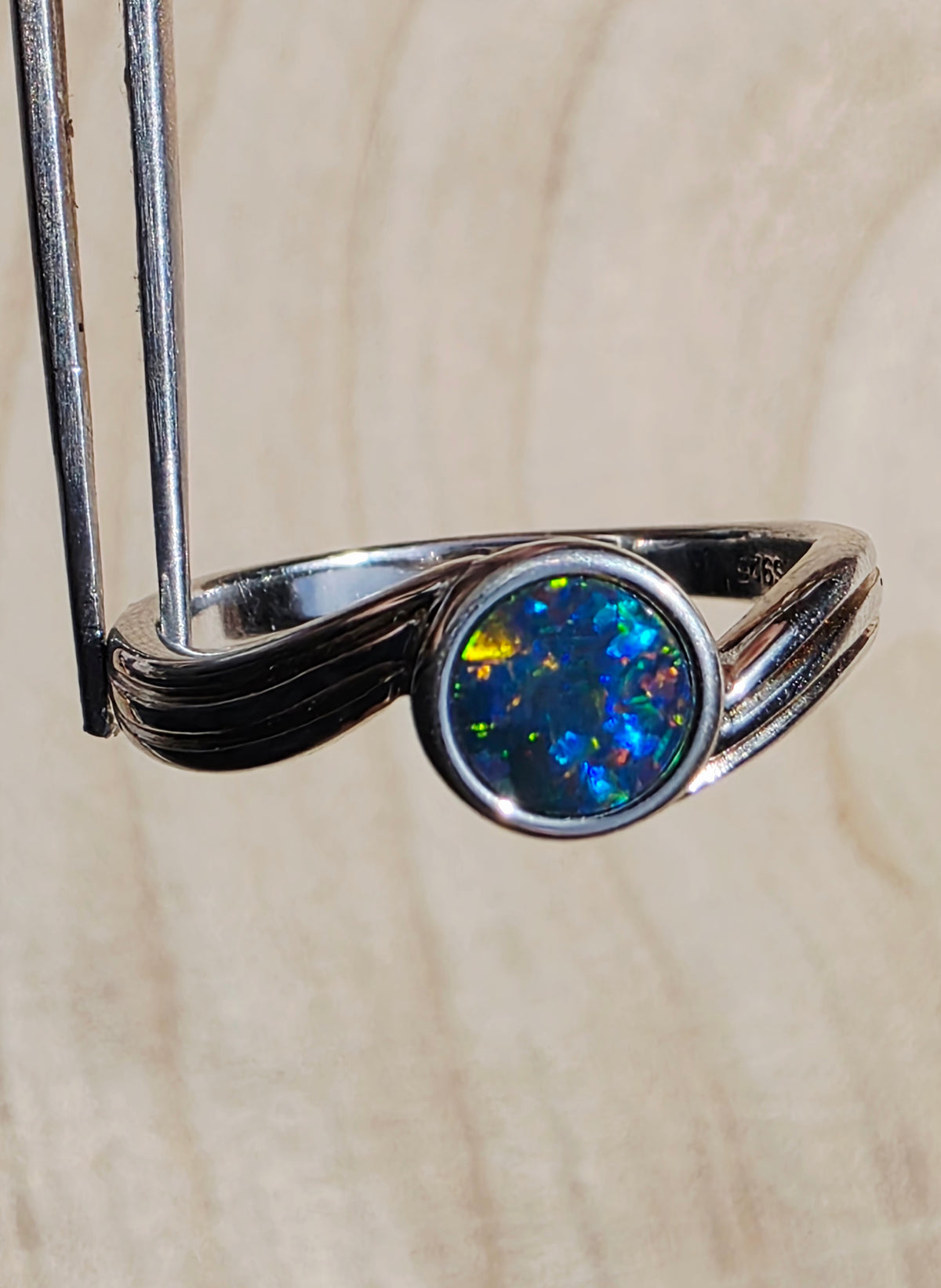 A silver ring with a round iridescent opal gemstone, held by a metal stand against a light wood background.